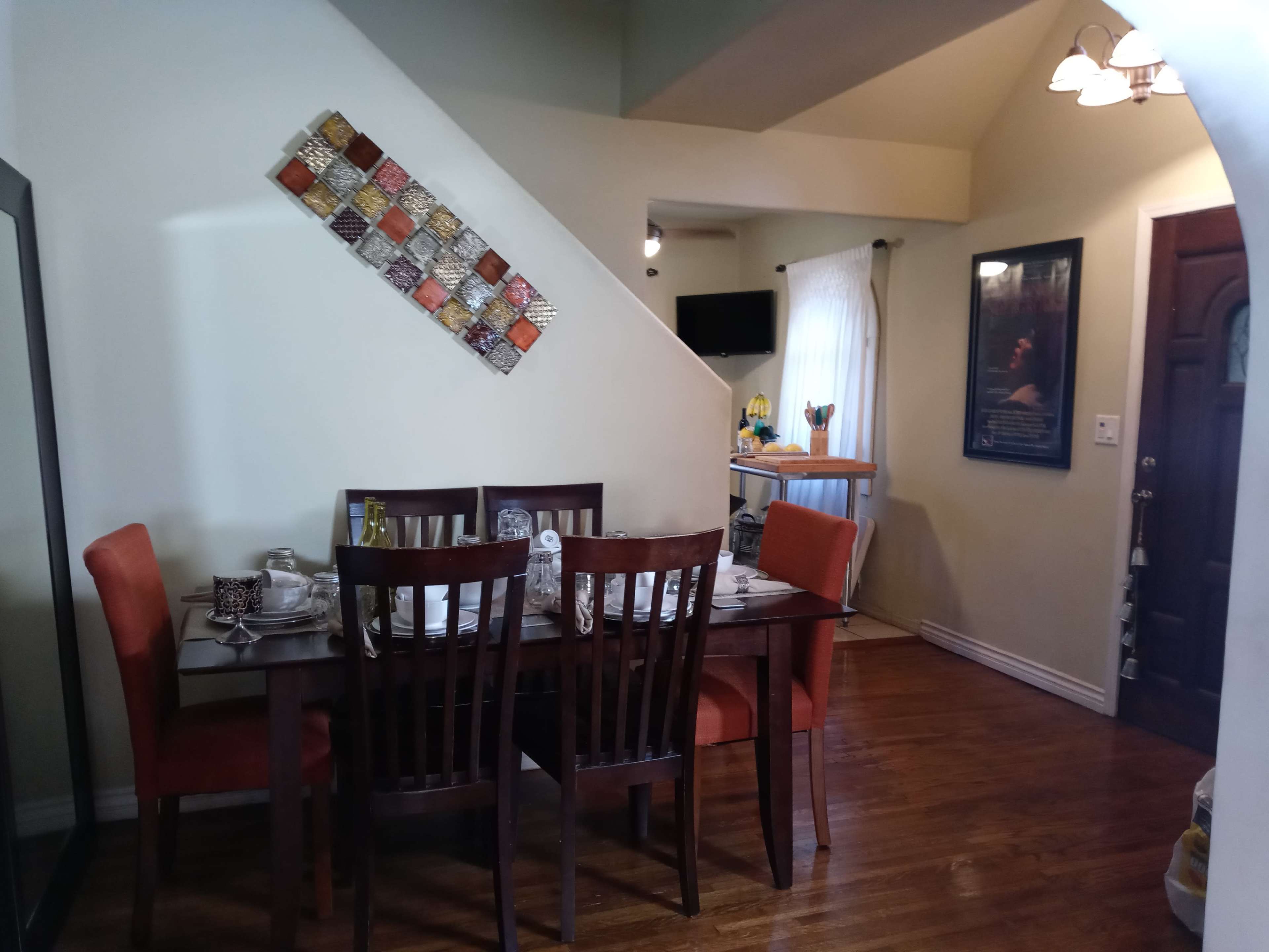 The image shows a dining area with a wooden table set for a meal, surrounded by chairs, and a decorative wall piece above the table.