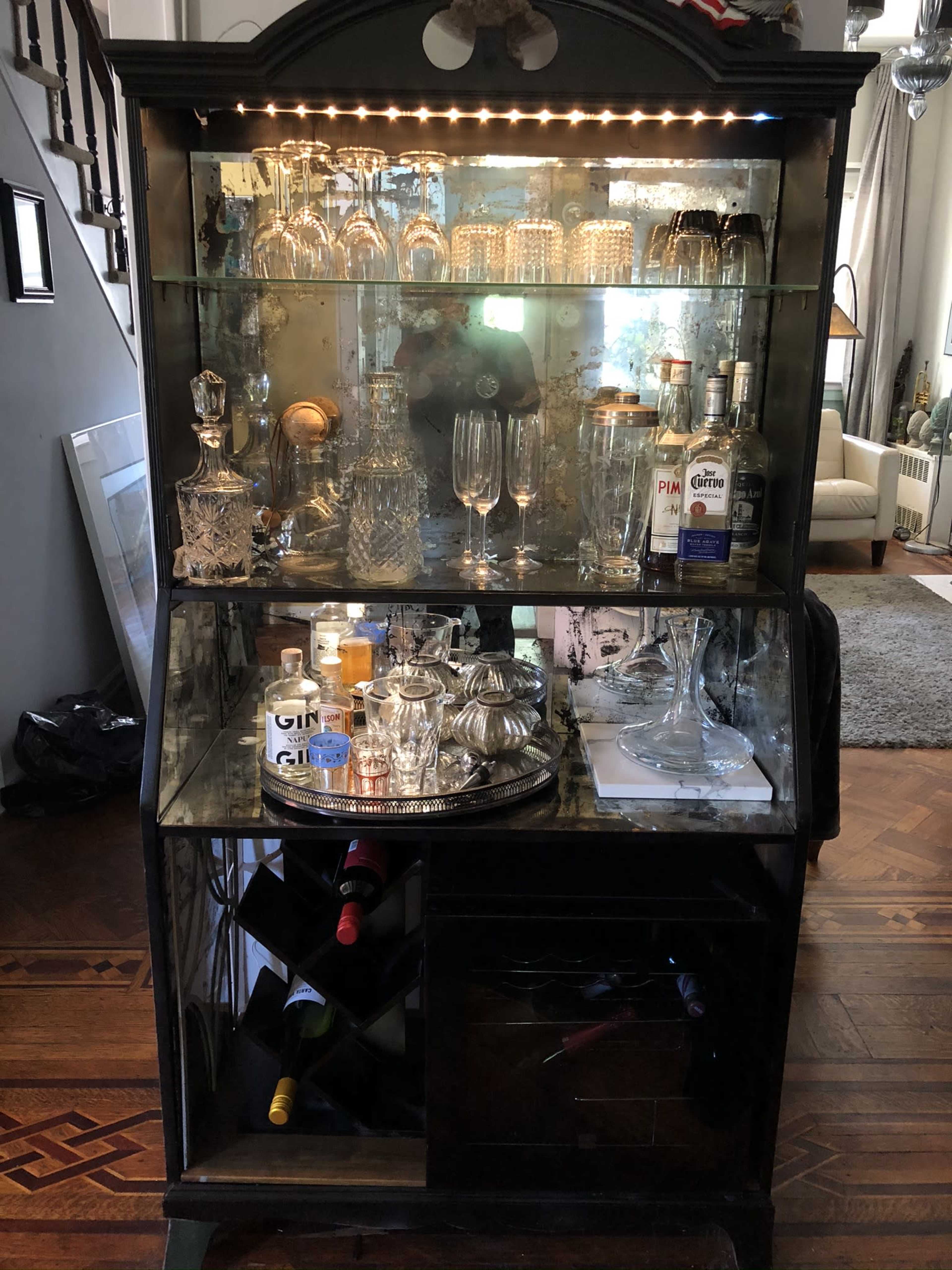 A mirrored bar cart displays an assortment of glassware, bottles, and a cocktail shaker, with a reflective back and shelves illuminated by lights.