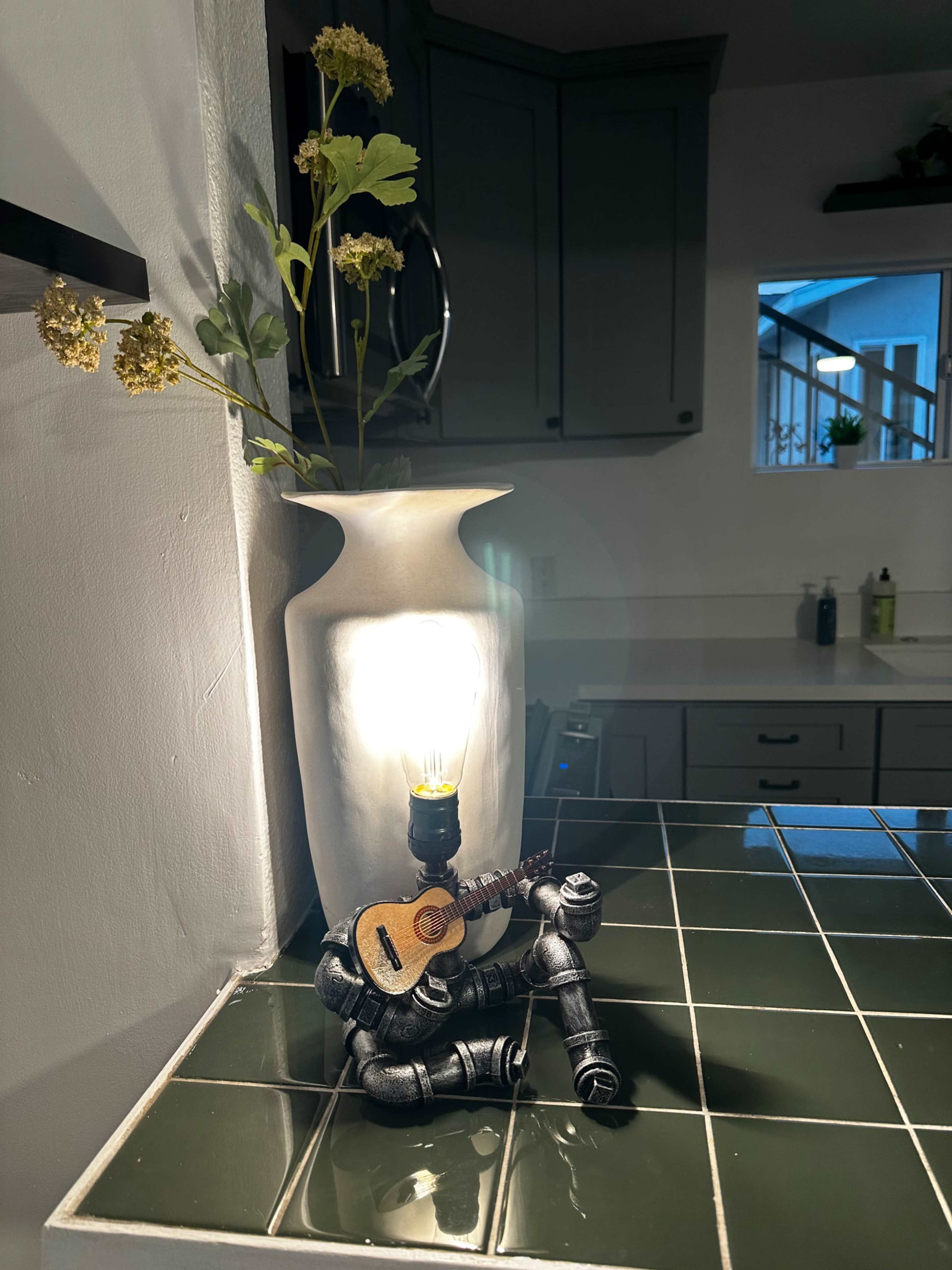 A decorative lamp shaped like a robot is sitting on a countertop, playing a small guitar beside a white vase with flowers.