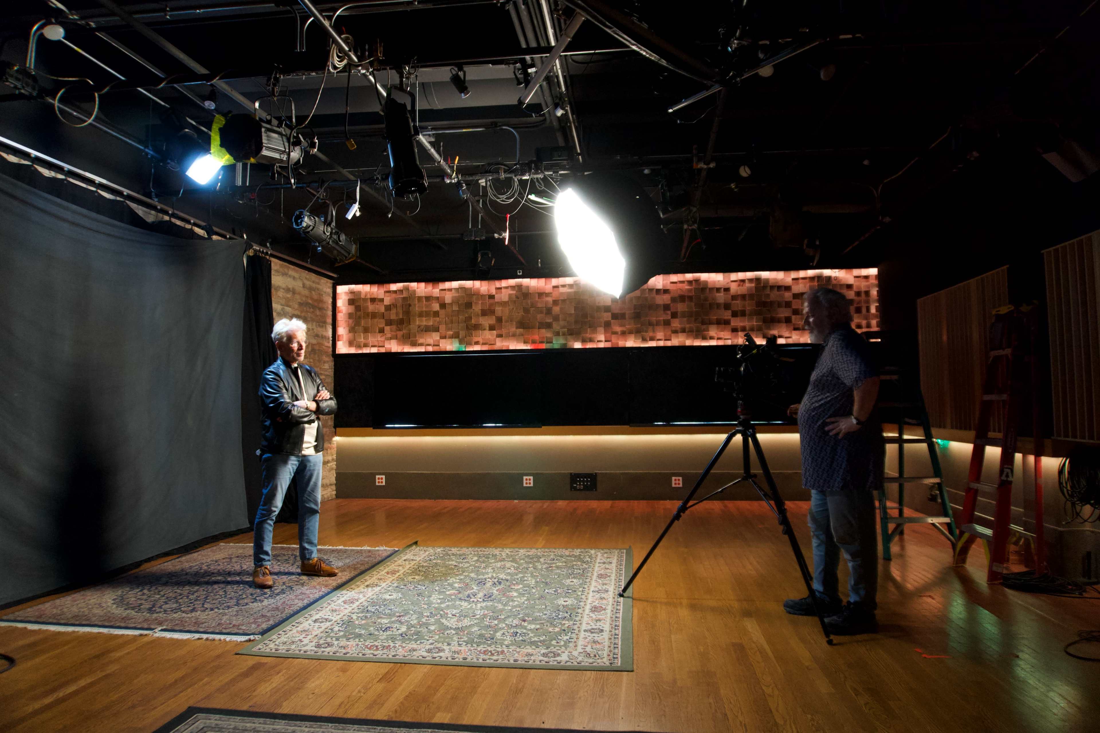 A photographer is setting up a shoot in a studio with a backdrop and lighting, while a man stands posed on an area rug.