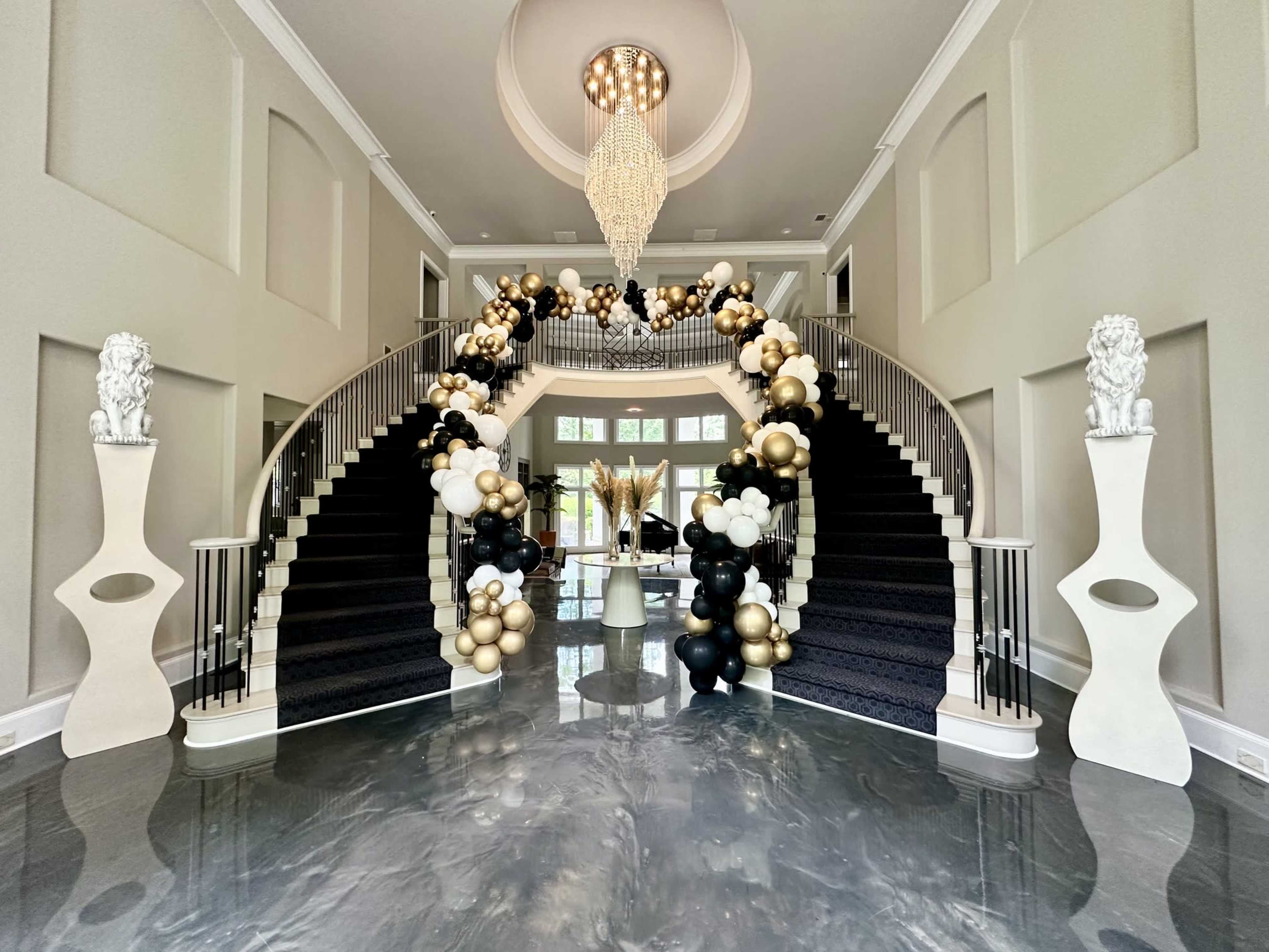 The image shows a spacious entryway featuring a grand staircase adorned with black, white, and gold balloon decorations, flanked by two sculptural columns with lion motifs.
