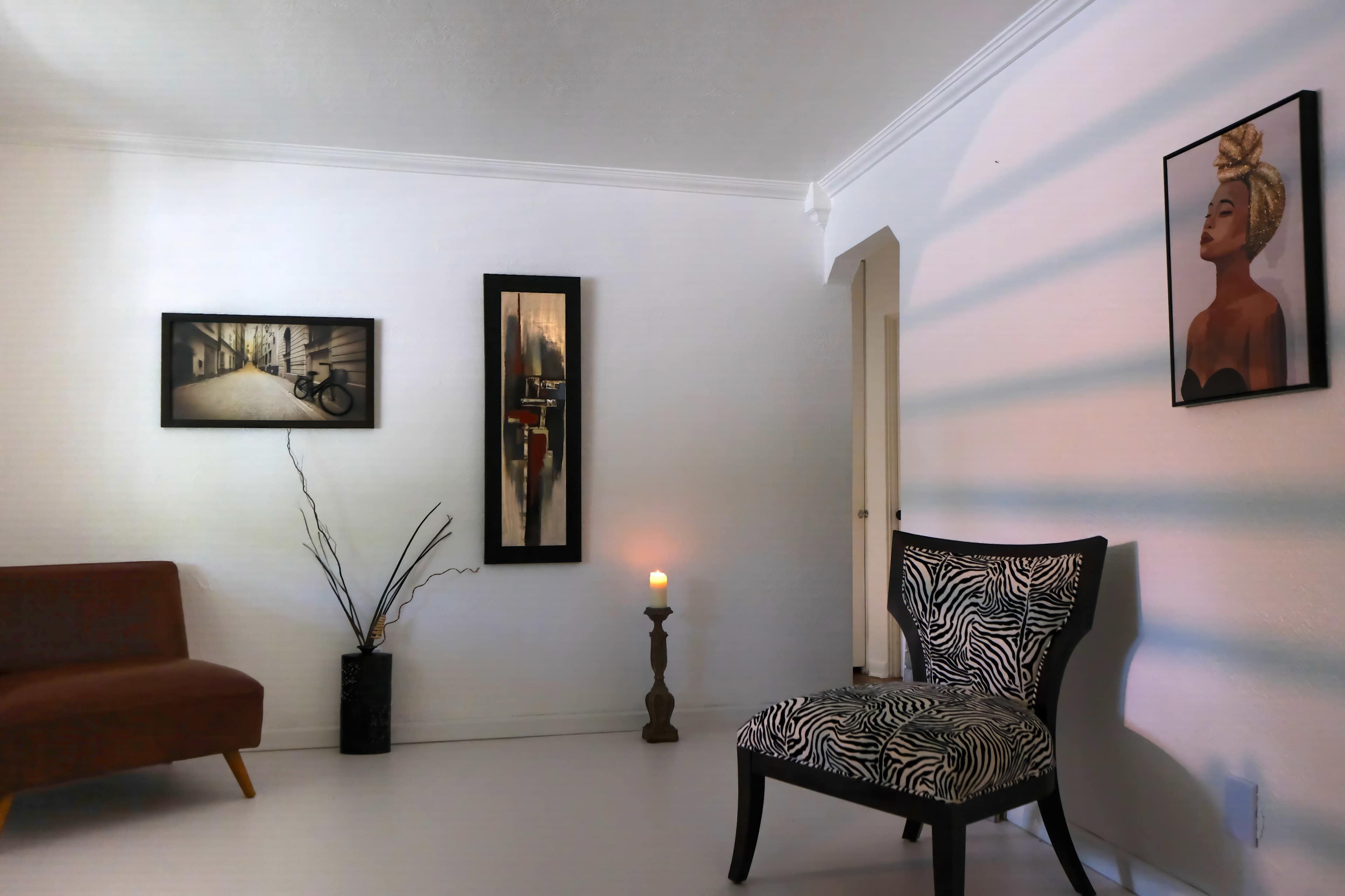 The image depicts a minimalist room with a zebra-patterned chair, a small wooden sofa, two framed artworks on the walls, a candle on a pedestal, and a decorative branch in a vase.