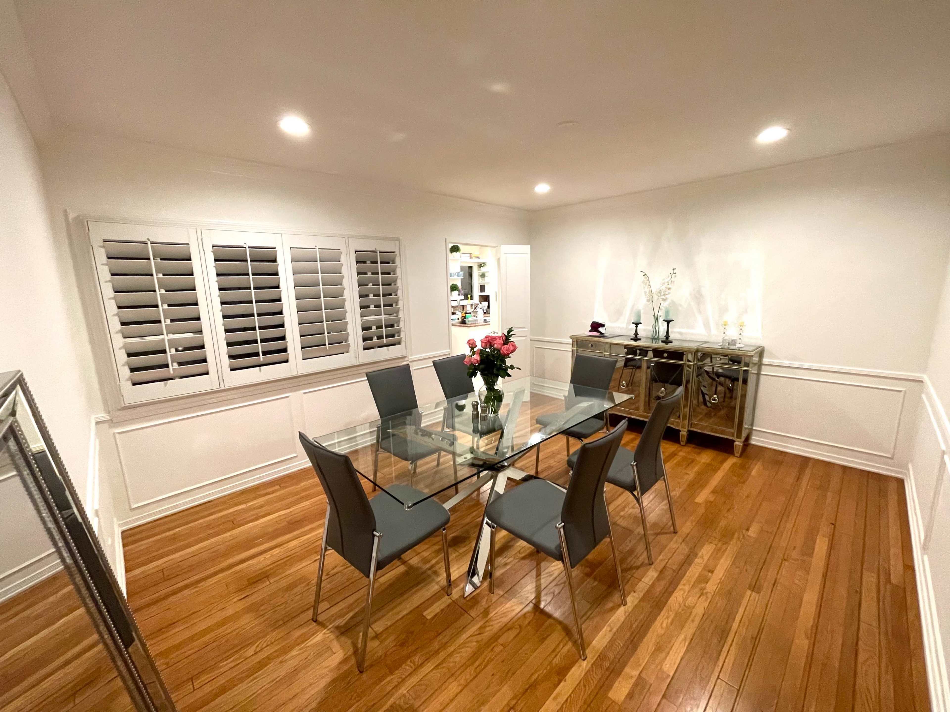 A glass dining table with six gray chairs is centered in a room featuring hardwood floors and white walls.
