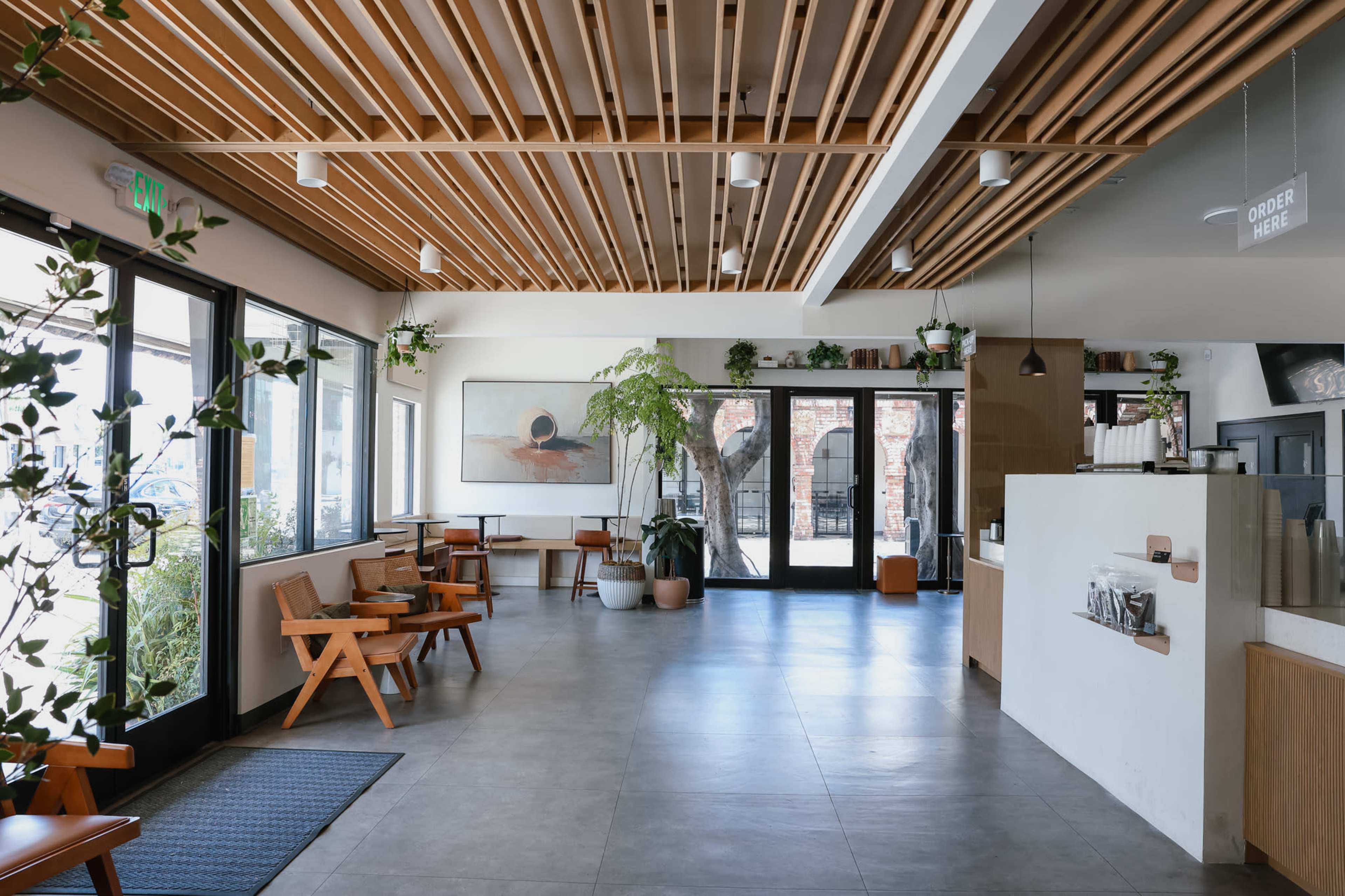 The image shows a bright, modern café interior featuring wooden ceiling beams, large windows, and a minimalist decor with seating areas and greenery.