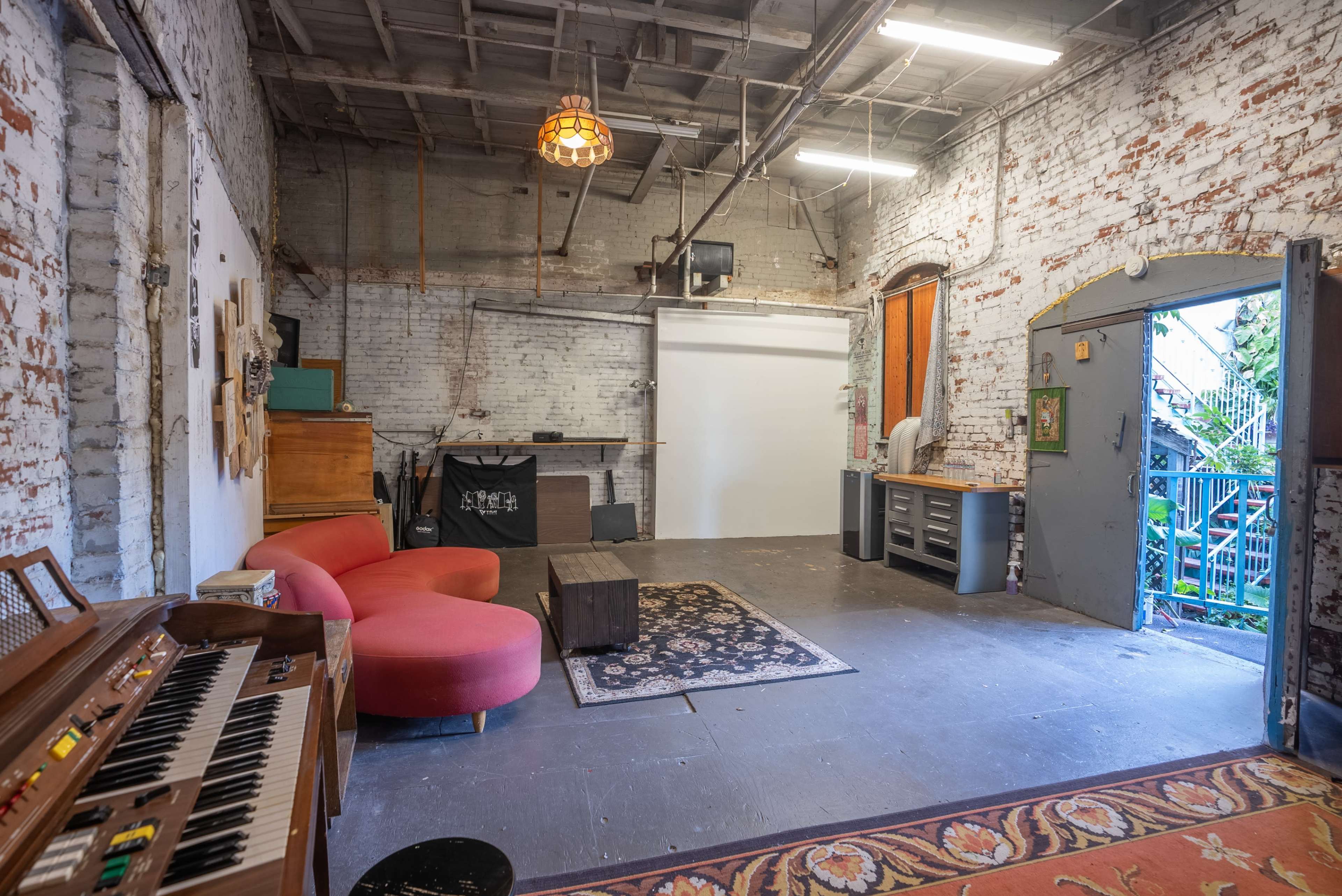 An industrial-style studio space with exposed brick walls, a red couch, a wooden piano, and a large white screen for presentations.