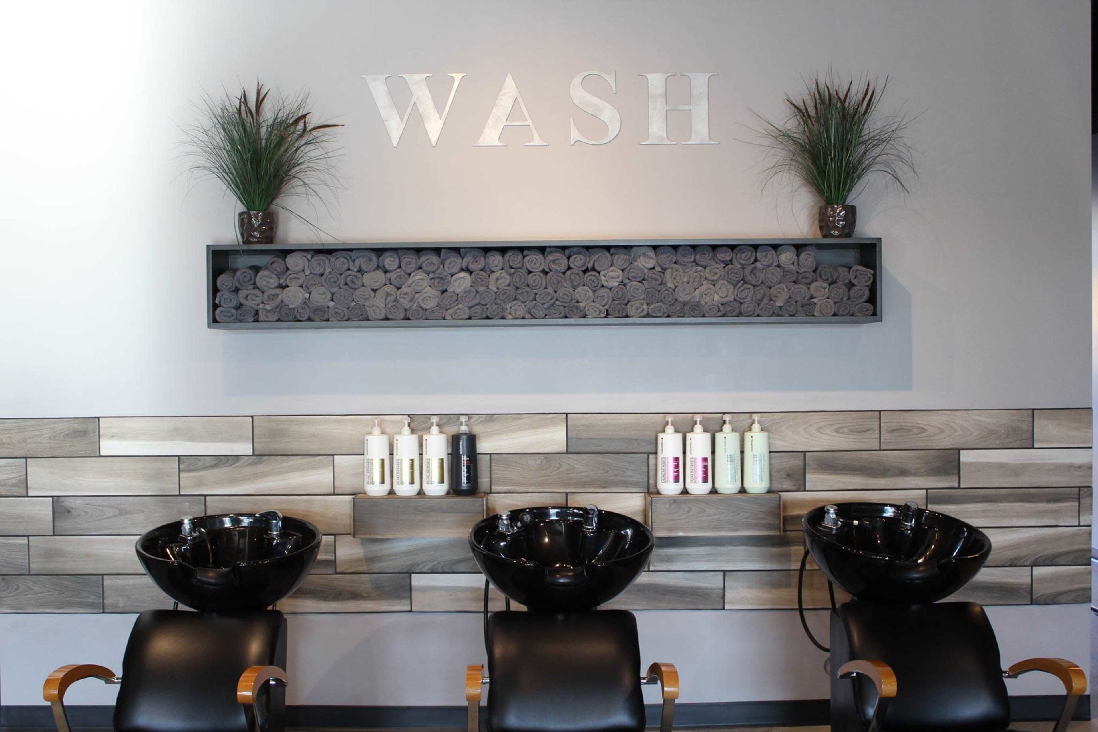 The image shows a hair salon wash area with three black shampoo bowls, a decorative shelf displaying rolled towels, and various hair care products lined up on a wooden shelf.
