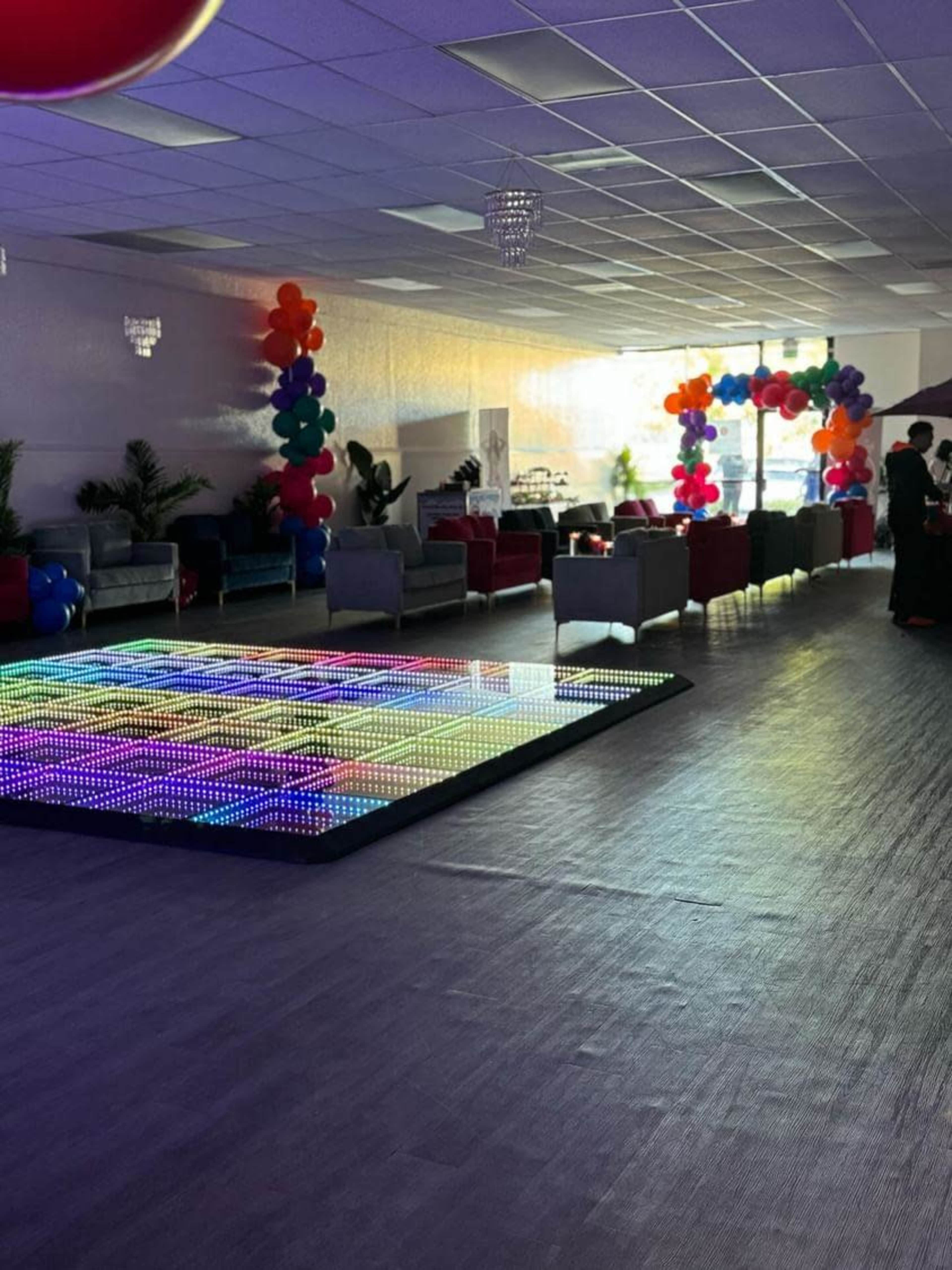 A spacious event venue with colorful balloon decorations, seating areas, and a light-up dance floor.