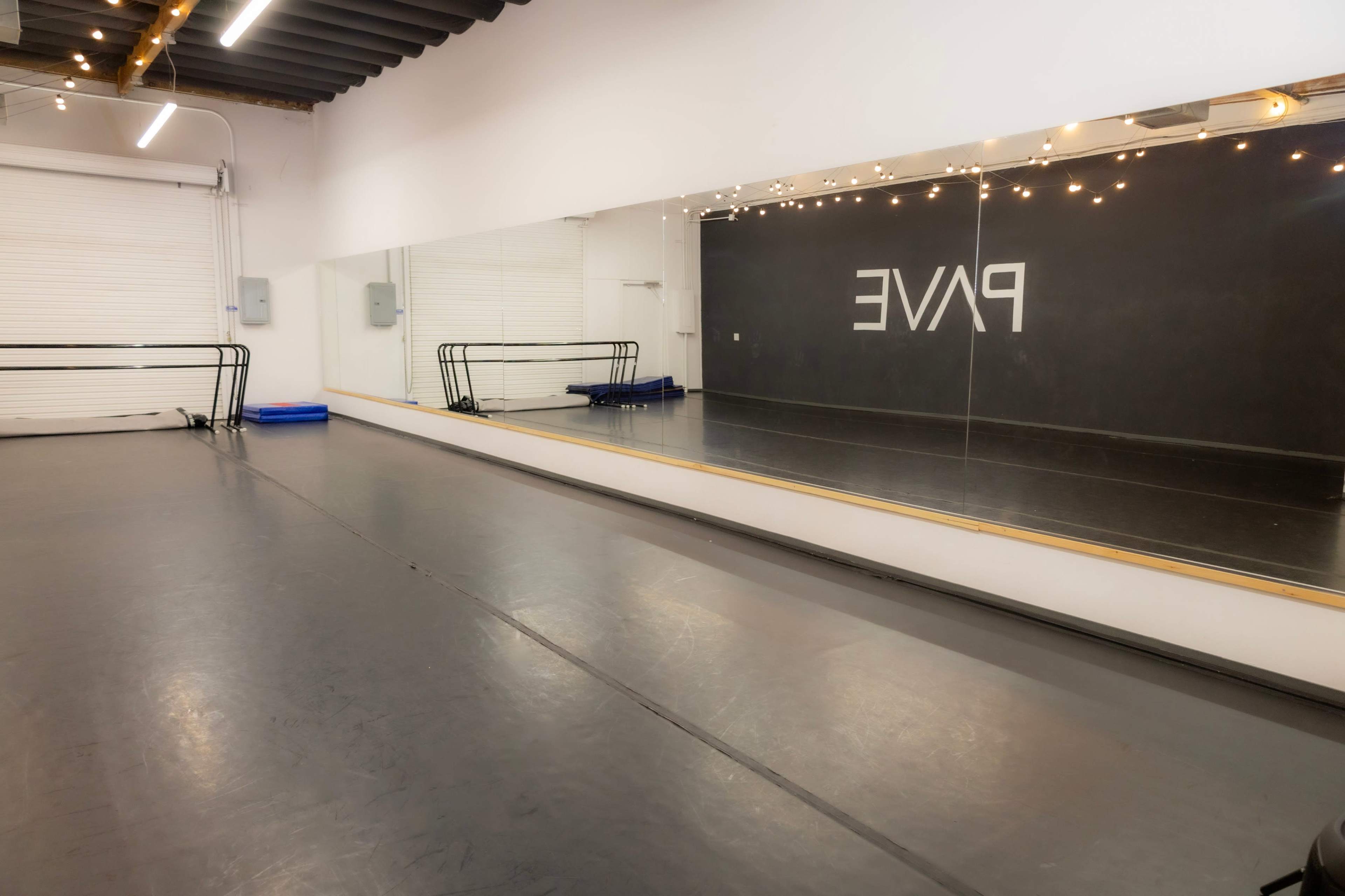 The image shows a spacious dance studio with a mirrored wall, a barre along one side, and a dark backdrop featuring the word "PAVE."