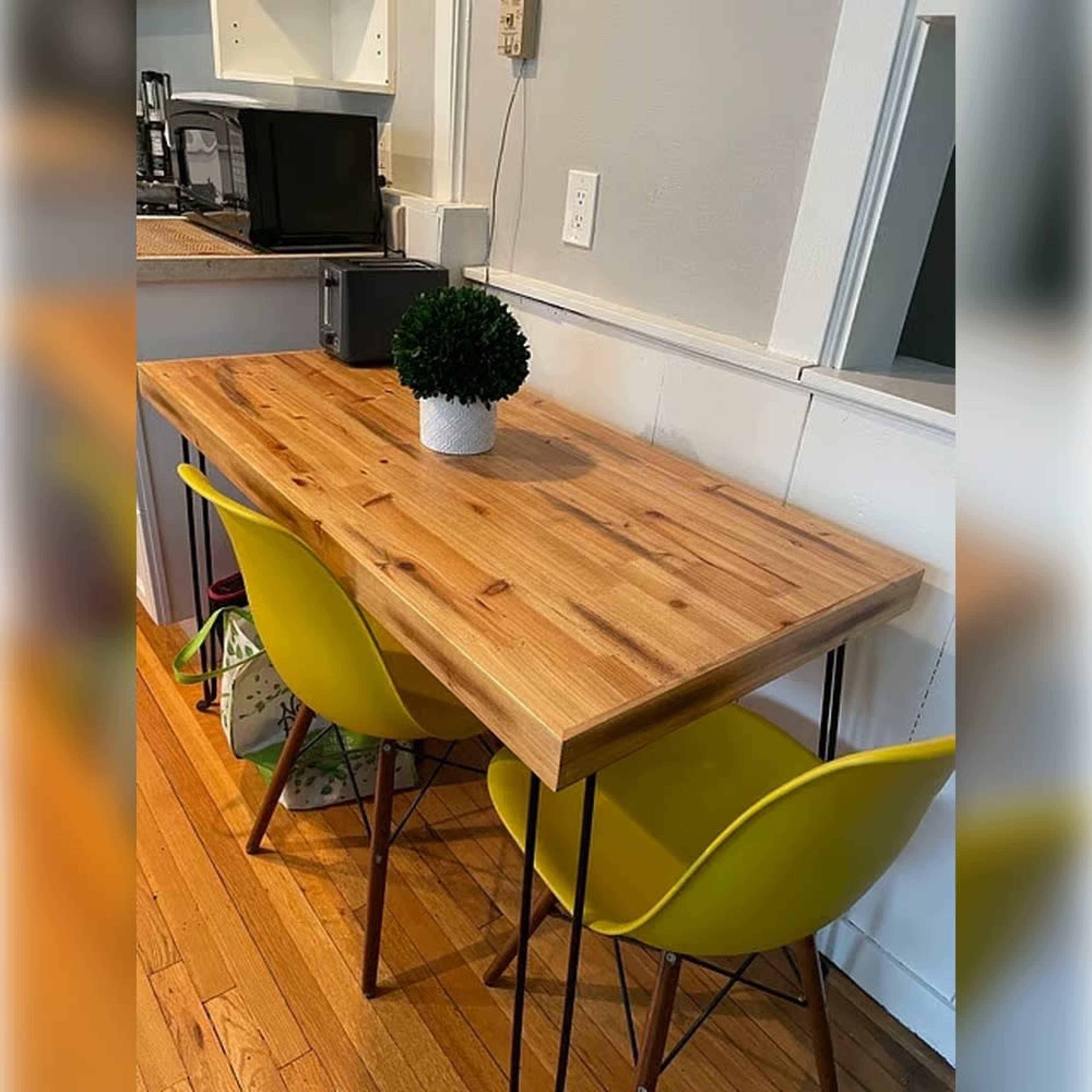 A wooden table with a smooth surface is set against a wall, accompanied by two yellow chairs and a small potted plant on top.