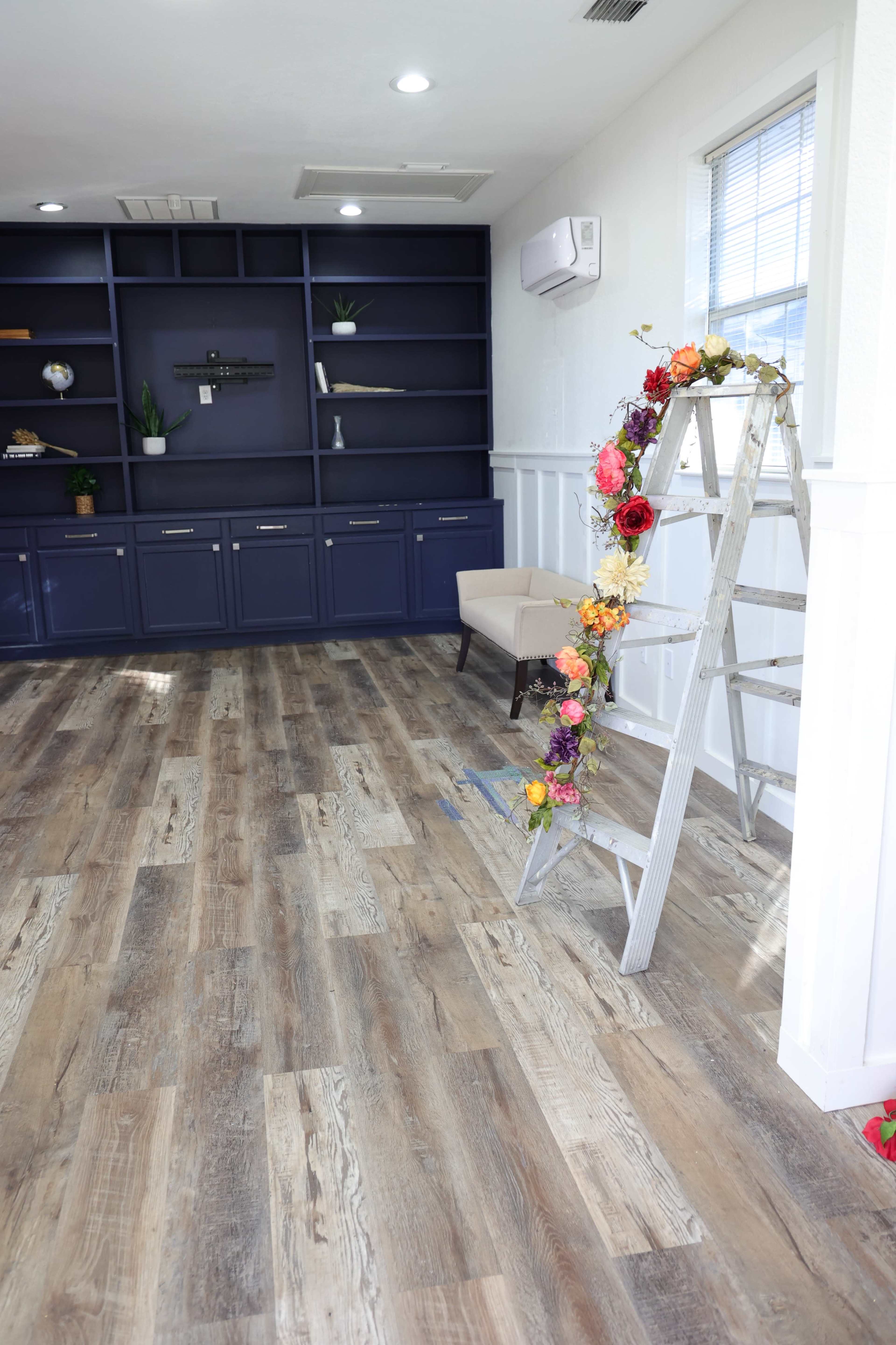 A ladder adorned with a floral garland stands next to a built-in shelving unit and a chair in a brightly lit room with wooden flooring.