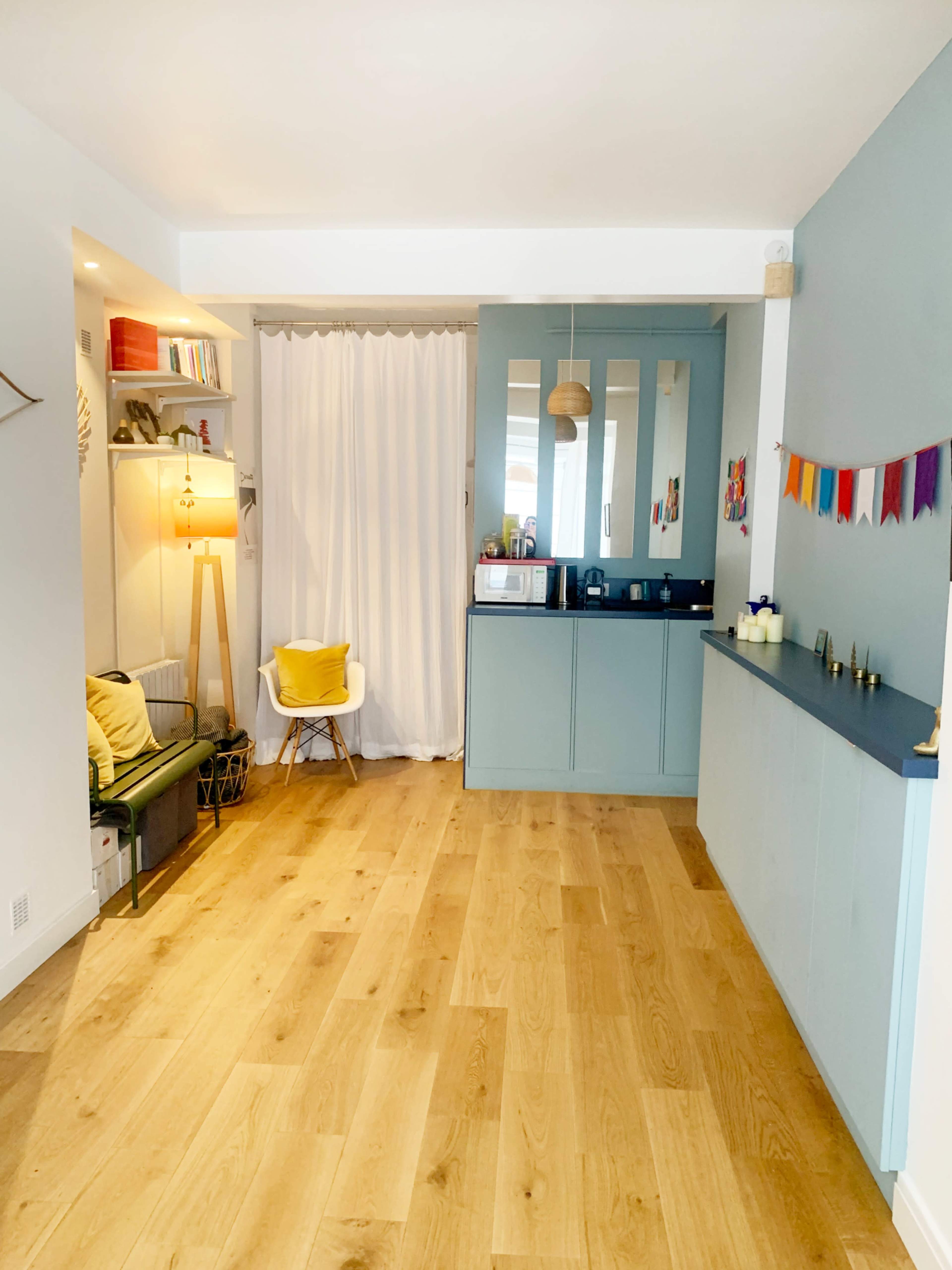A bright room features light wooden flooring, a kitchenette with teal cabinetry, a small seating area, and a curtain dividing the space.