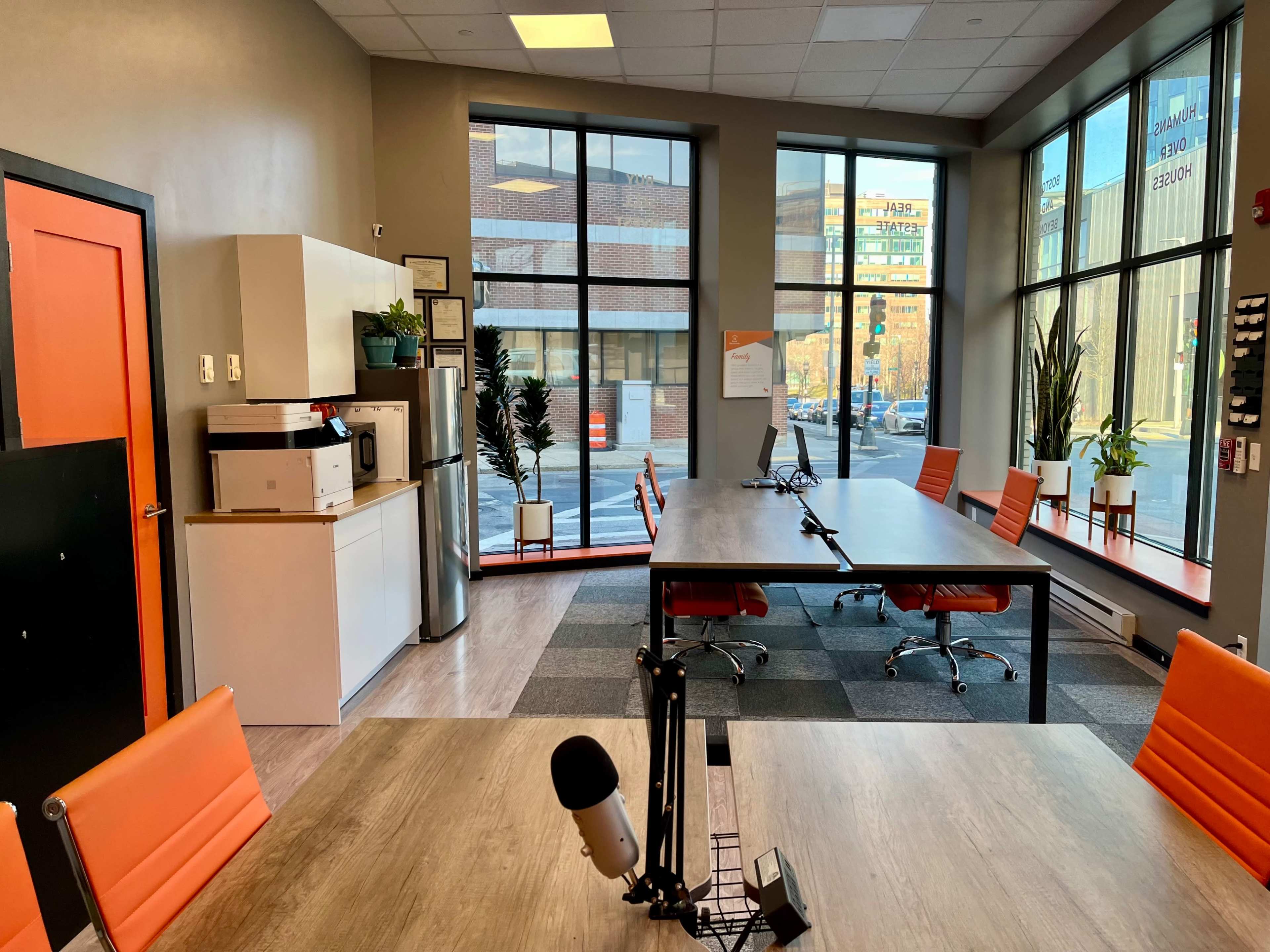 The image shows a modern office space with a large table, orange chairs, and large windows allowing natural light to enter.