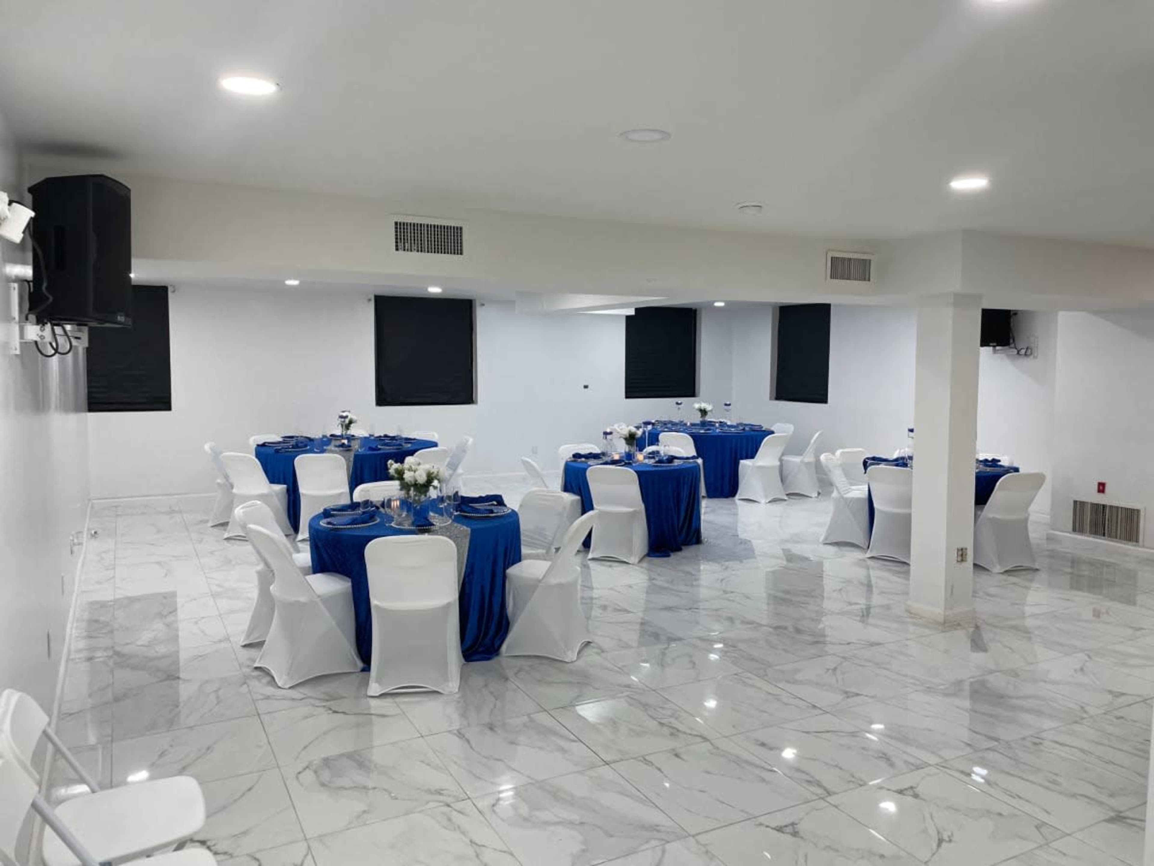The image shows a banquet hall with round tables covered in blue tablecloths and white chairs arranged neatly on a marble floor.