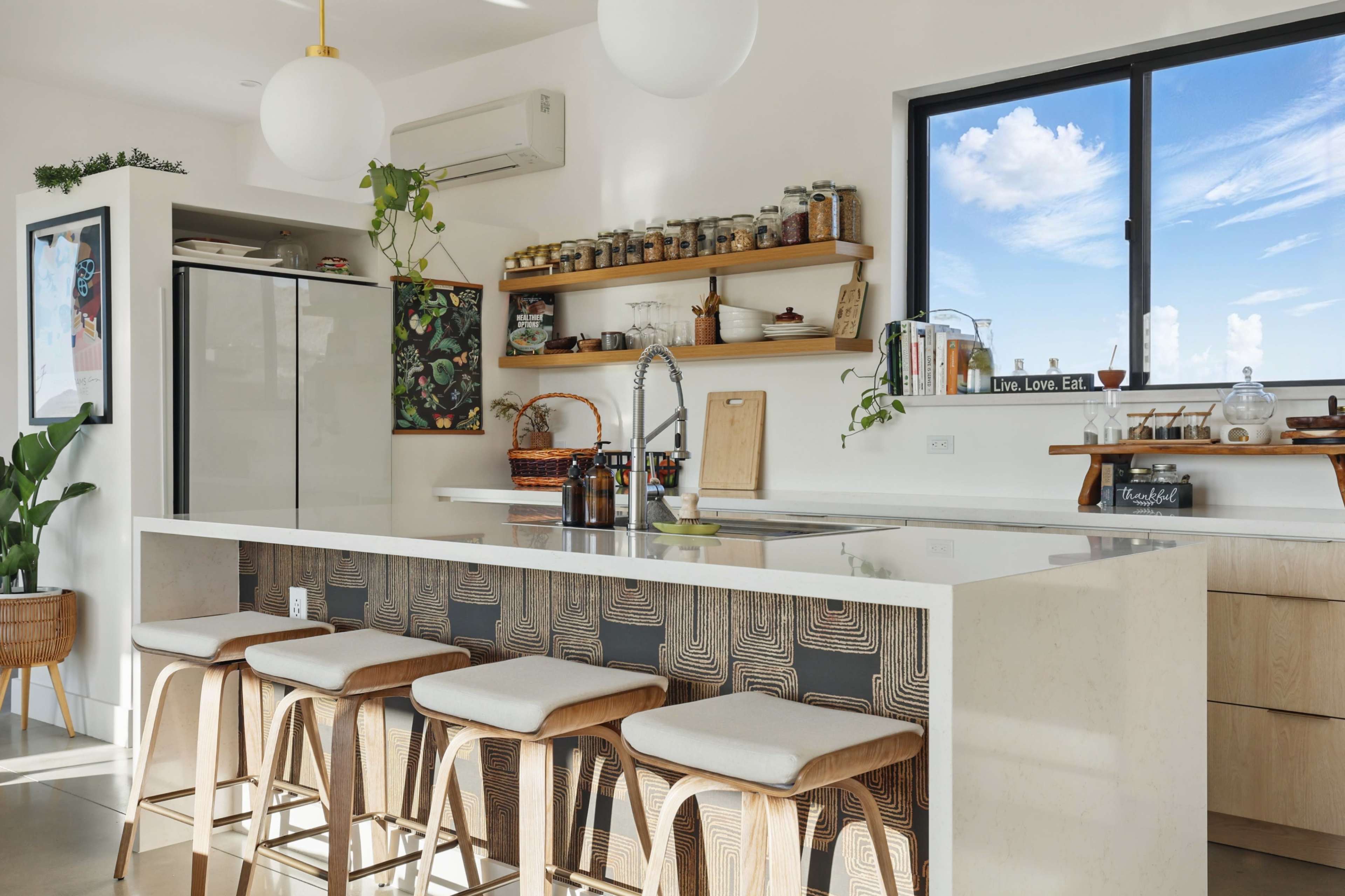 The image shows a modern kitchen with a large island, bar stools, open shelving, and a window displaying a clear sky.