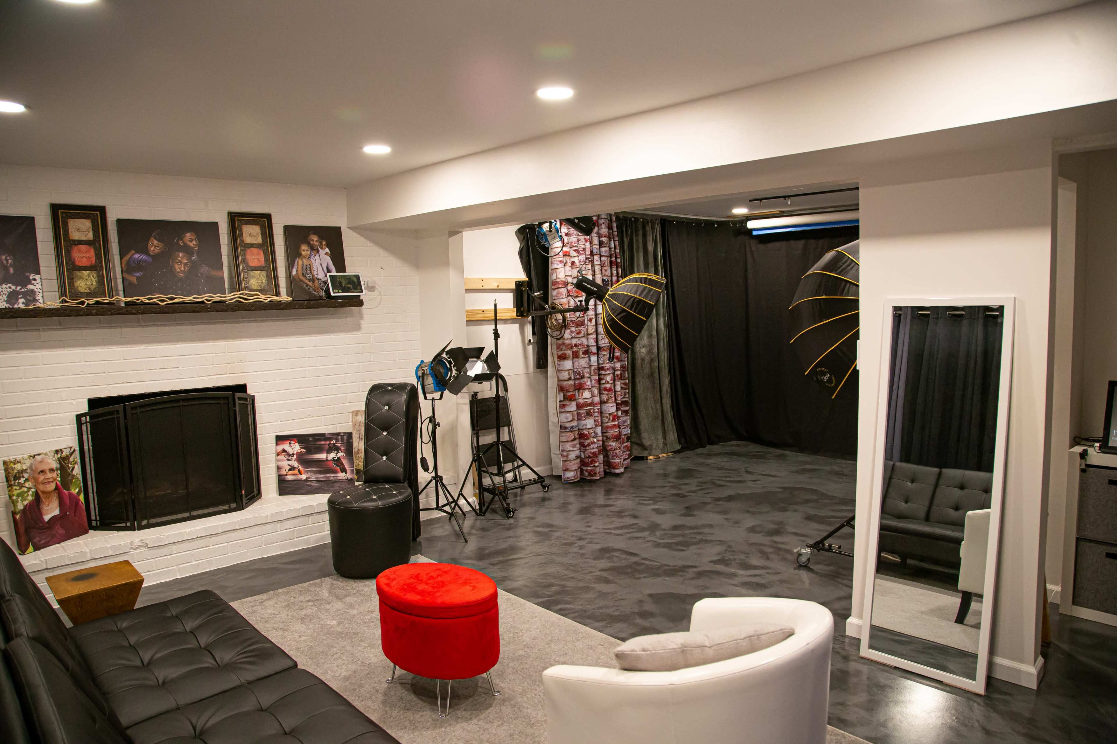 A studio space with a black backdrop, lighting equipment, a fireplace, and a seating area featuring a black couch and a red ottoman.