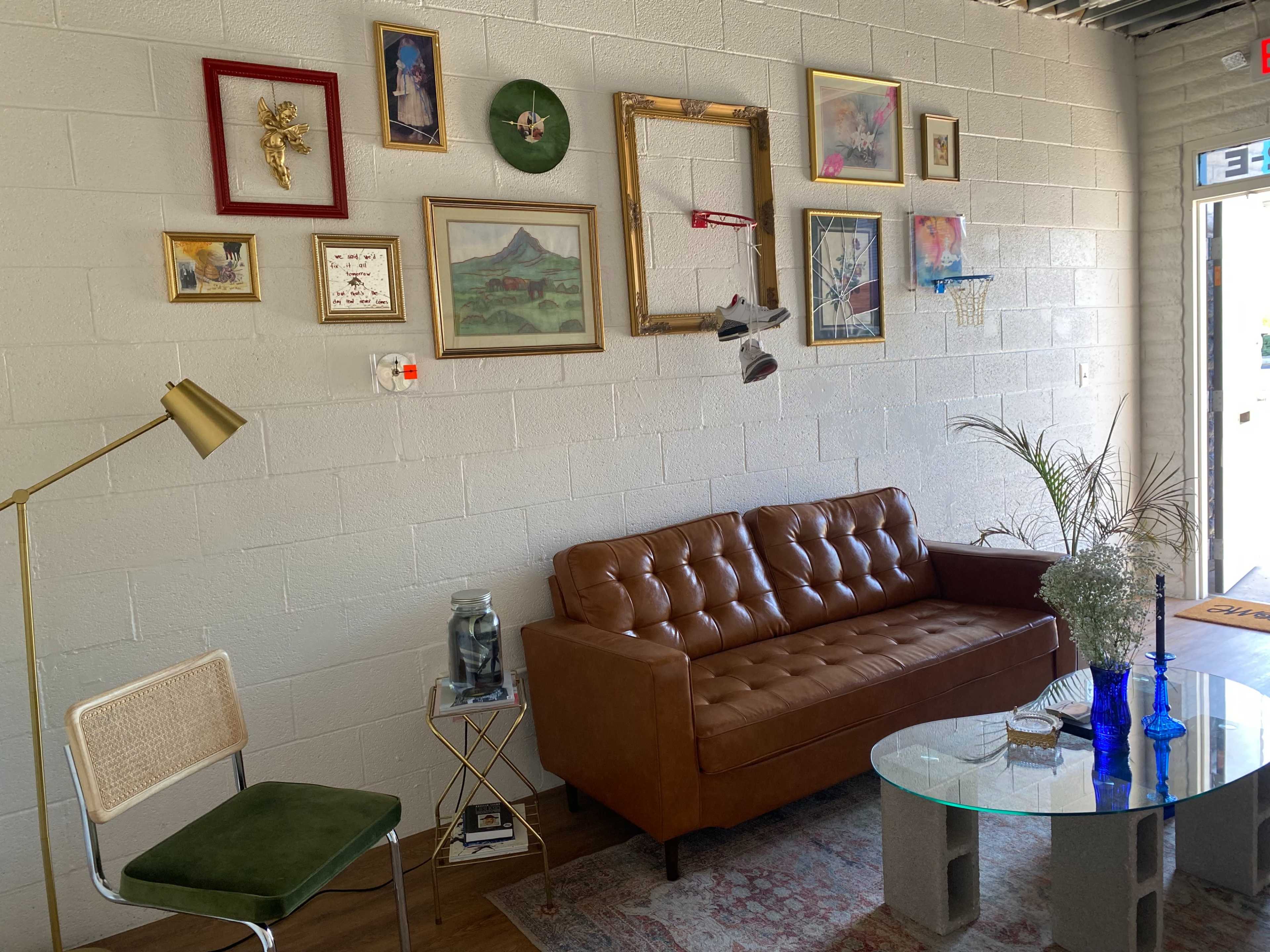 The image shows a cozy living area featuring a brown leather sofa, a glass coffee table, a green chair, and a wall decorated with various framed artworks and pictures.