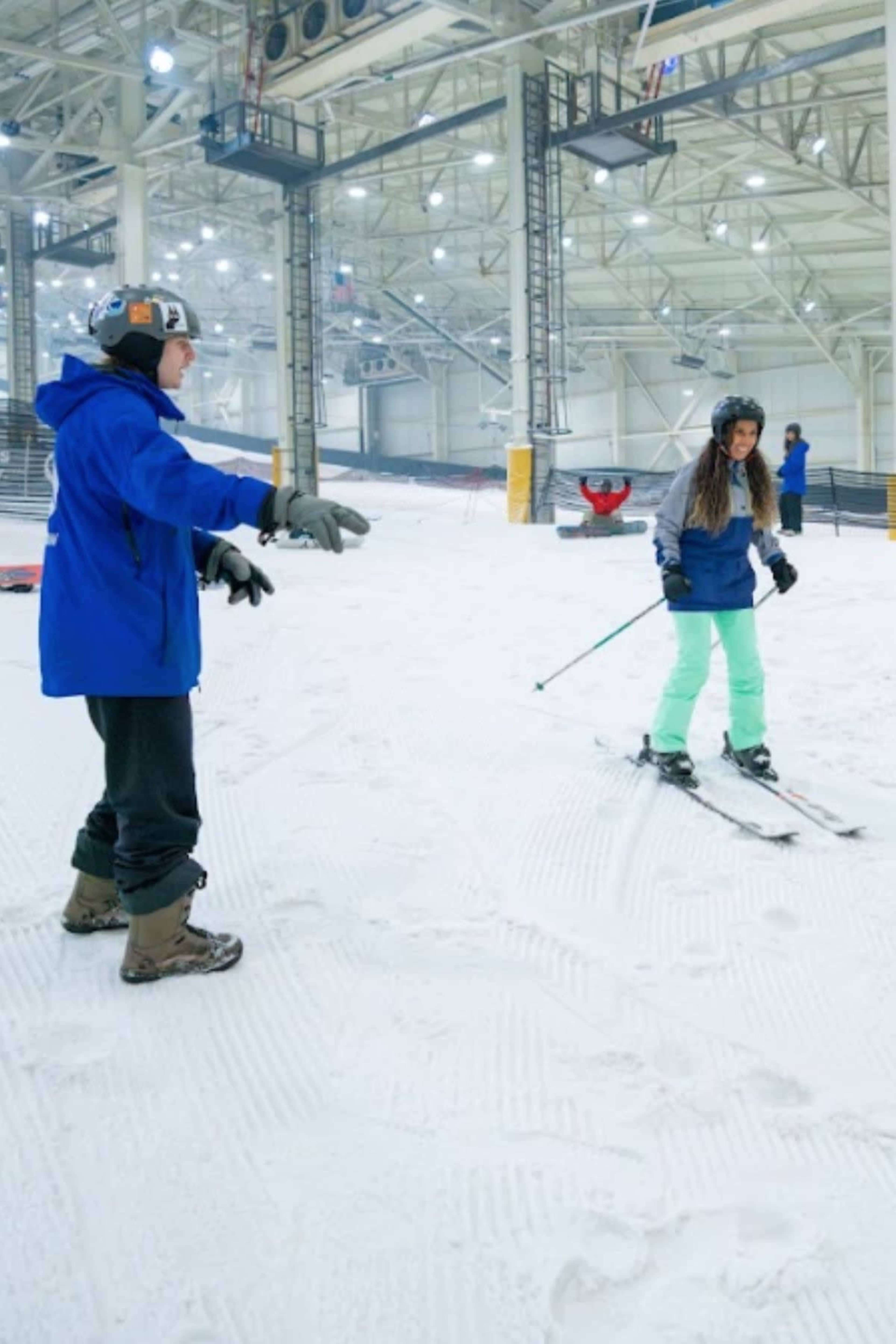 A ski instructor demonstrates techniques to a student on a snowy indoor slope.