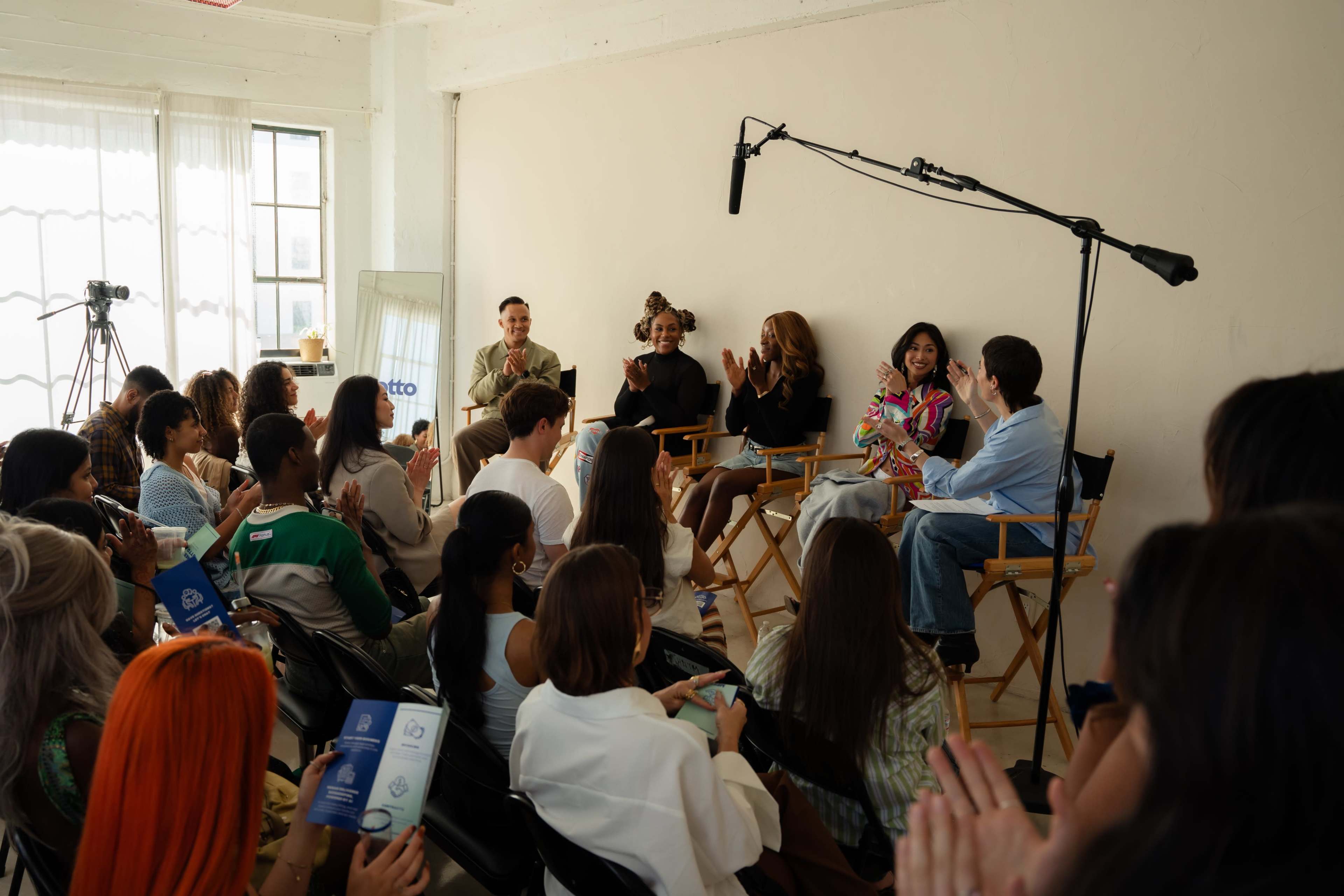 A diverse audience is seated in a bright studio, attentively watching as a panel discussion takes place on a stage with three speakers.