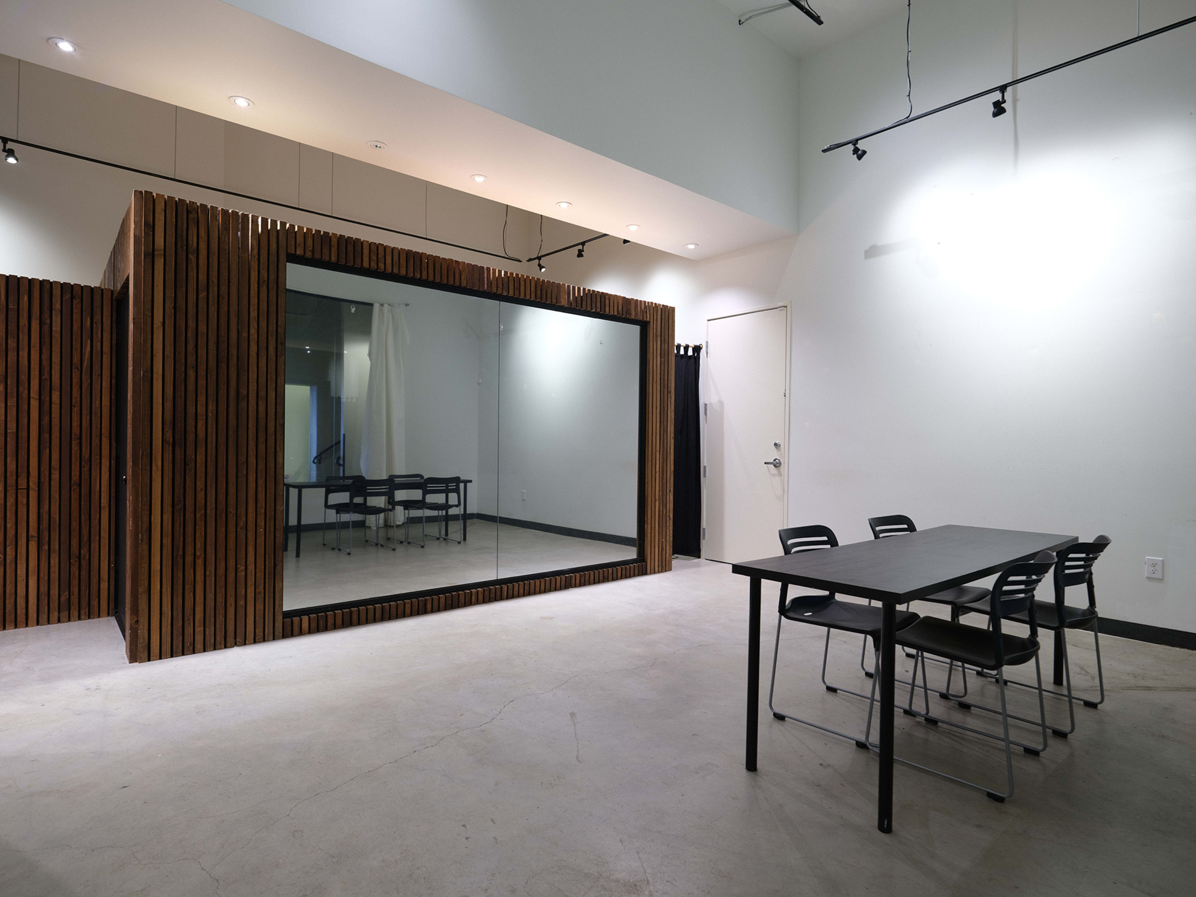 The image shows a modern room featuring a large mirror, wooden paneling, a black table, and several chairs arranged around it.