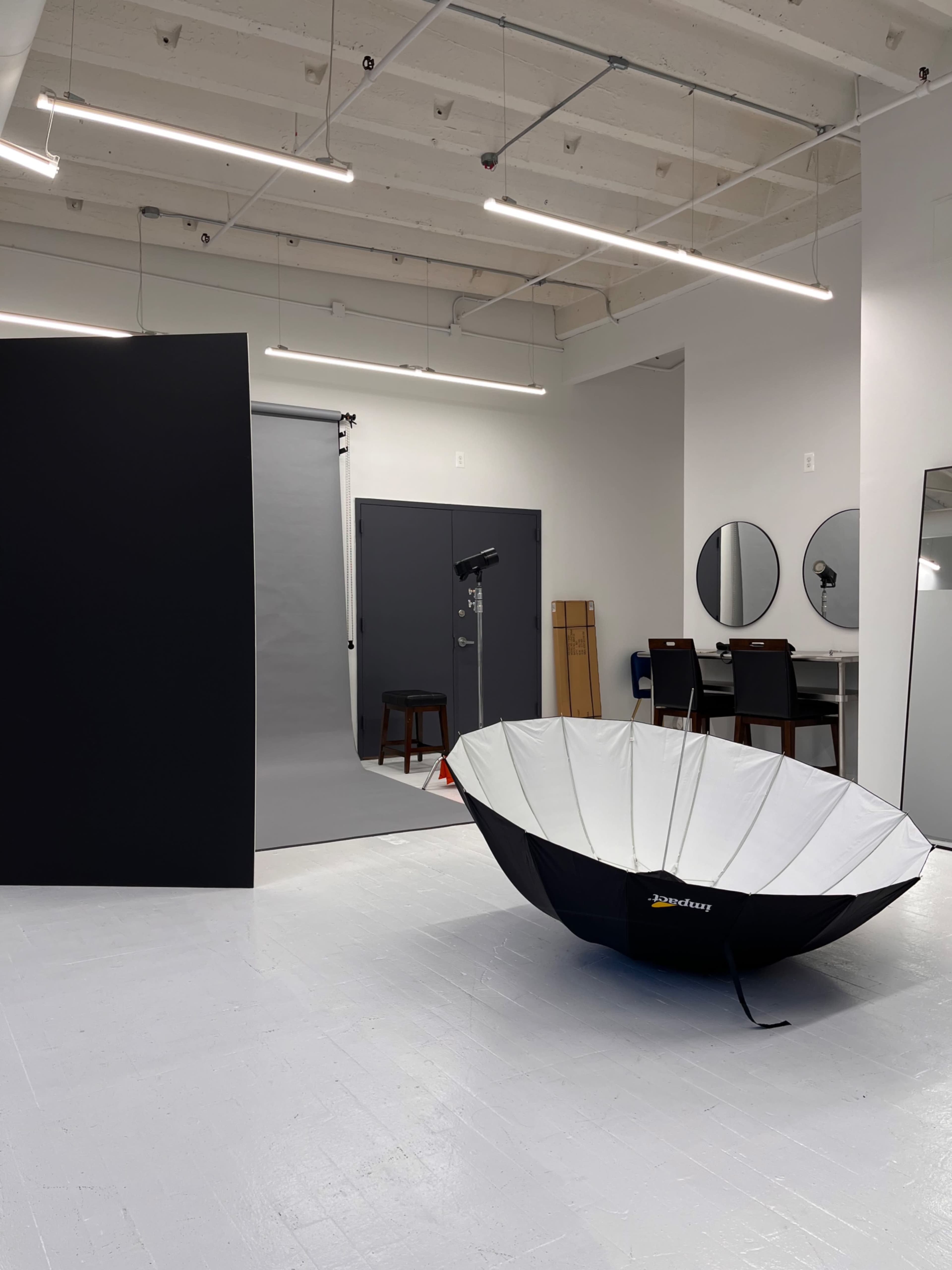 A large, black and white reflecting umbrella stands in a well-lit photography studio with a gray backdrop and several mirrors.