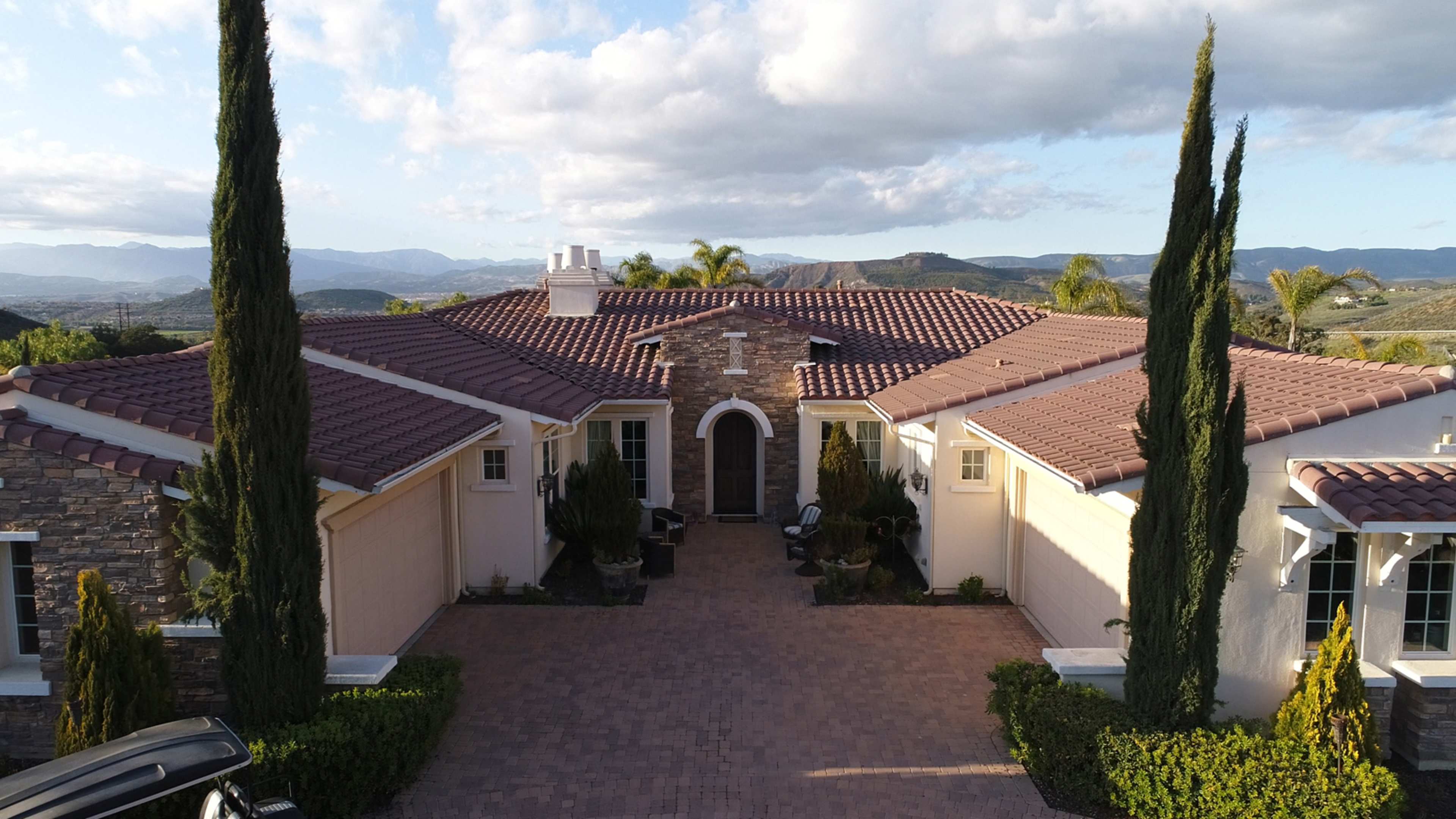 The image shows a large Mediterranean-style home with a stone façade, a tiled roof, and lush landscaping set against a mountainous backdrop.