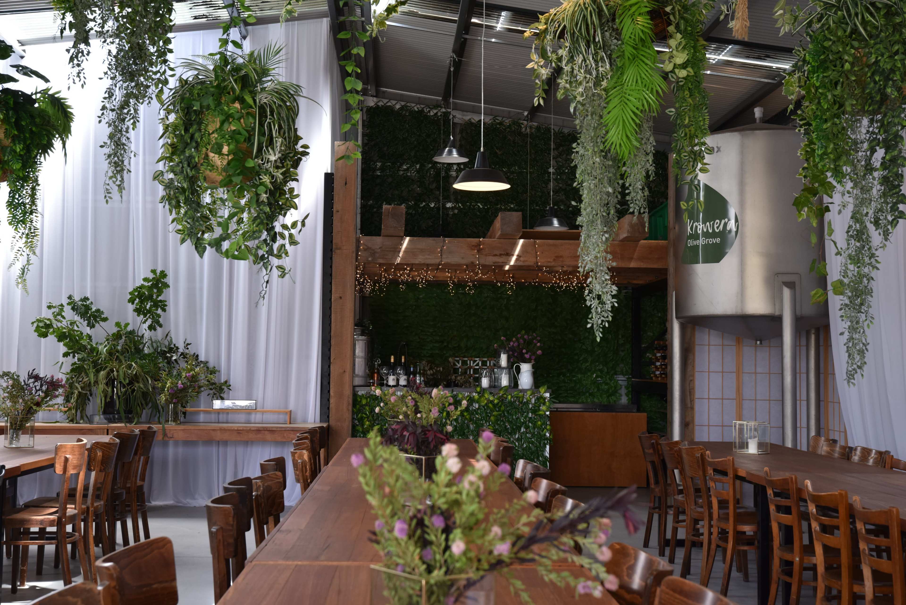 A spacious indoor dining area features long wooden tables surrounded by chairs, with greenery hanging from the ceiling and a bar at the back against a plant-covered wall.
