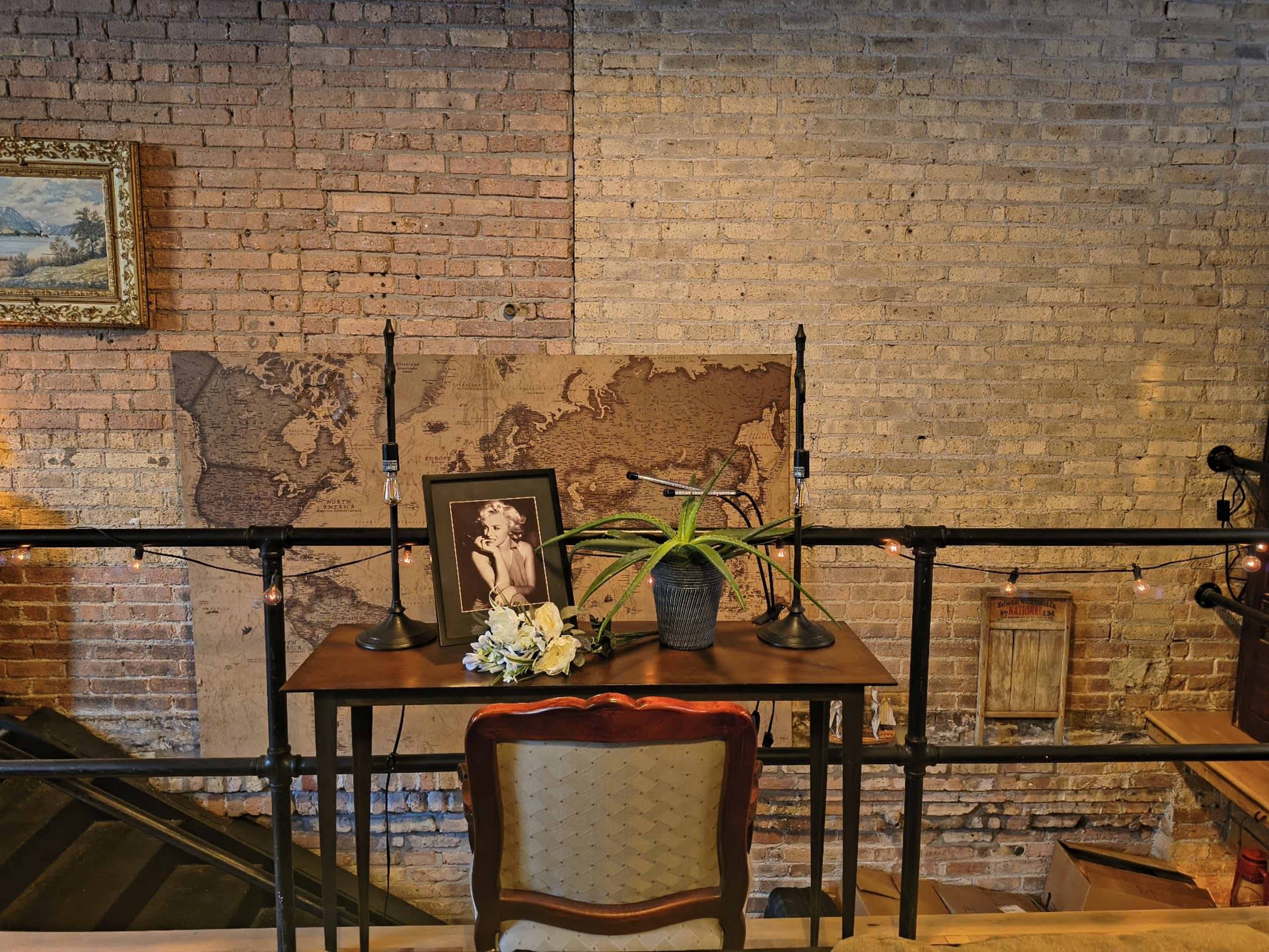 The scene features a wooden table with two lamps and a small potted plant, set against a brick wall decorated with a large world map and a vintage framed portrait.