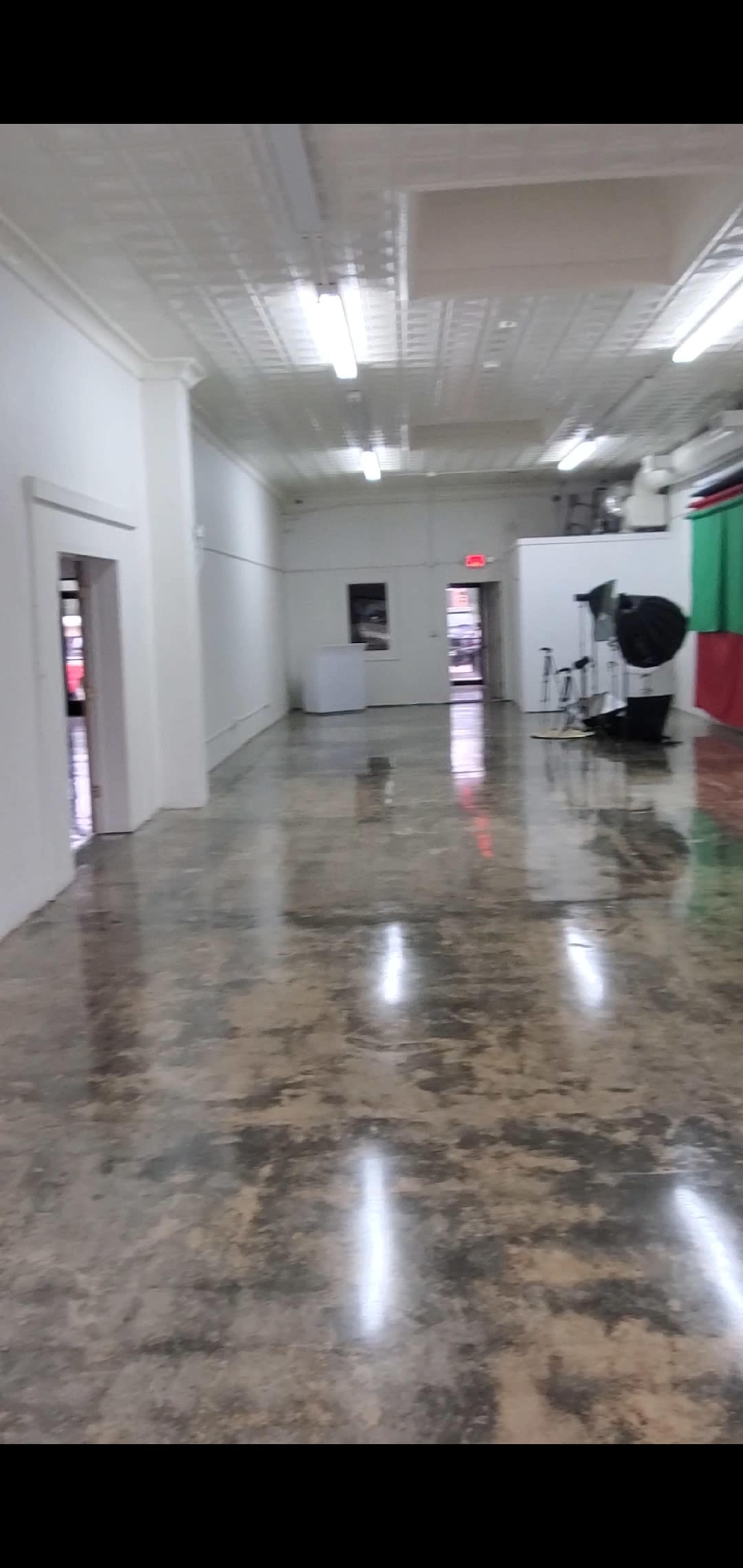 The image shows a spacious, empty hallway with polished concrete flooring and white walls, featuring a few doors and photography equipment in the background.