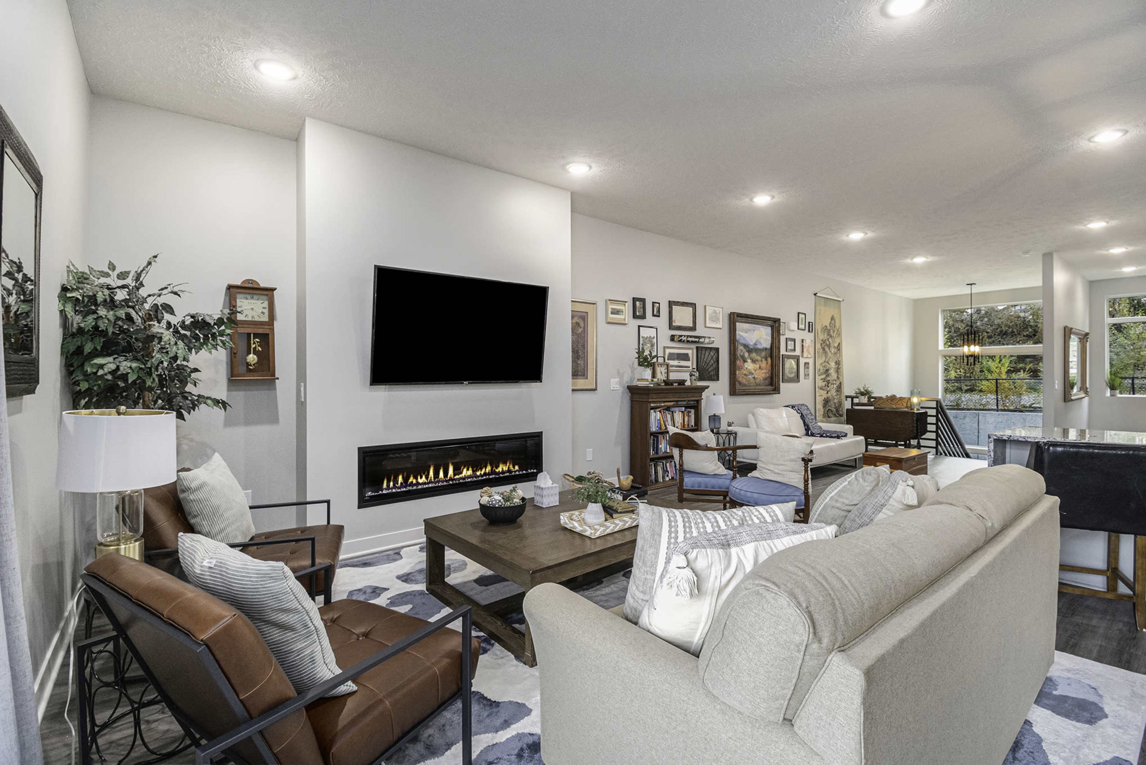 A modern living room features a flat-screen TV mounted on the wall, a fireplace underneath, and various pieces of furniture including a sofa, chairs, and a coffee table, all surrounded by framed artworks.
