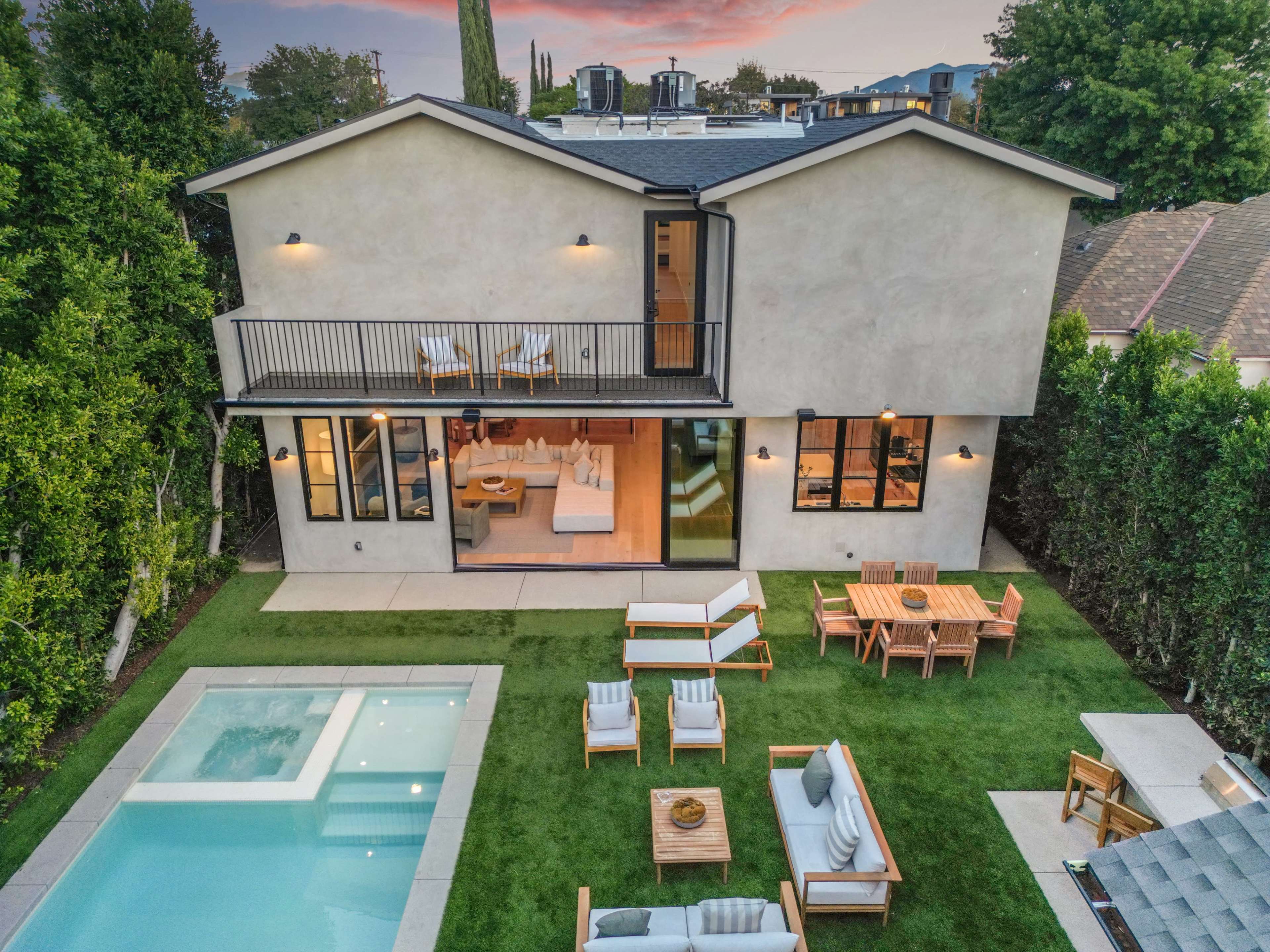 The image shows a modern two-story house with a swimming pool and outdoor seating area amidst well-maintained greenery.
