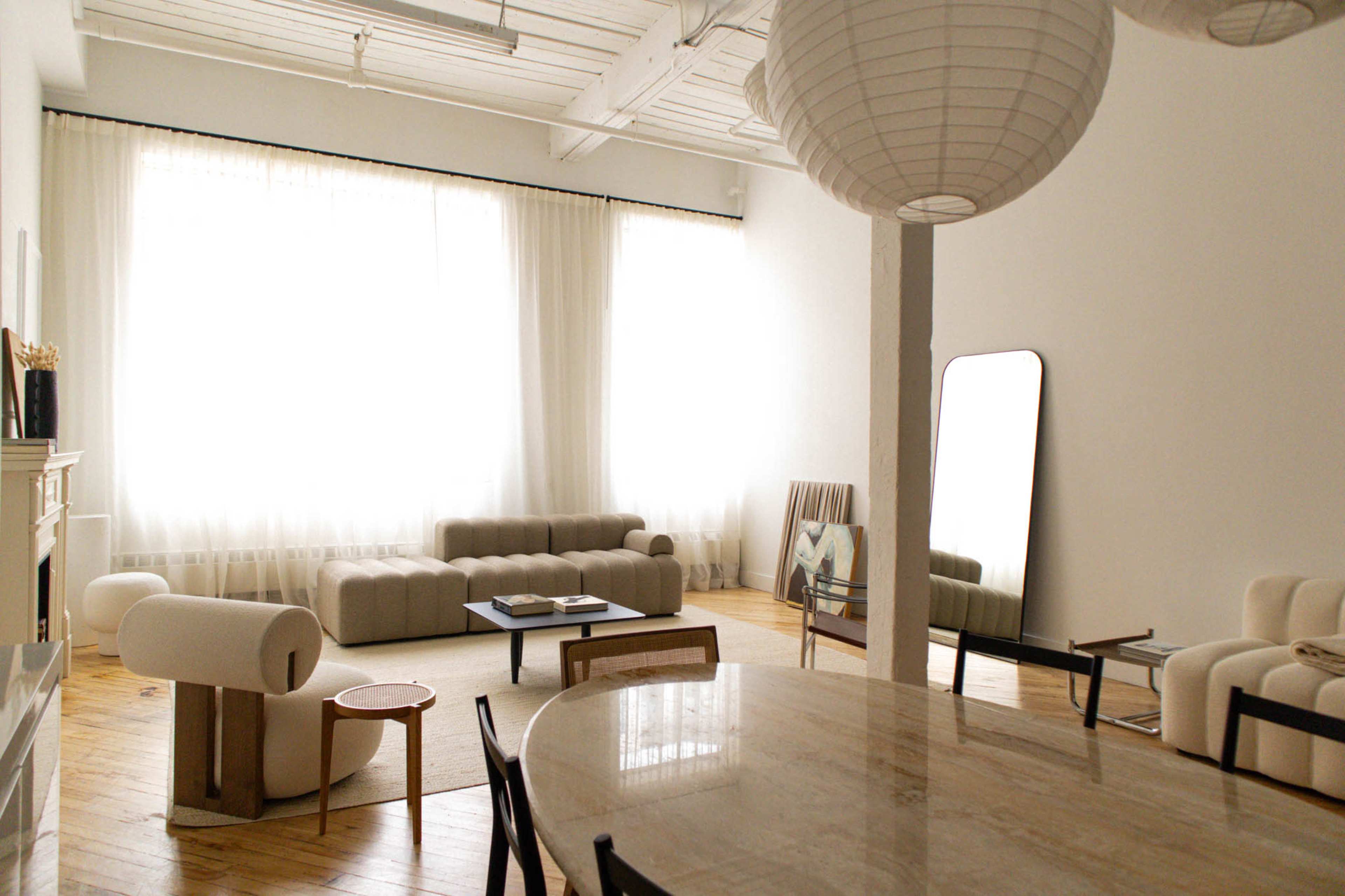 A spacious living area features large windows with sheer curtains, a sectional sofa, a mirror, a dining table, and minimalist furniture.