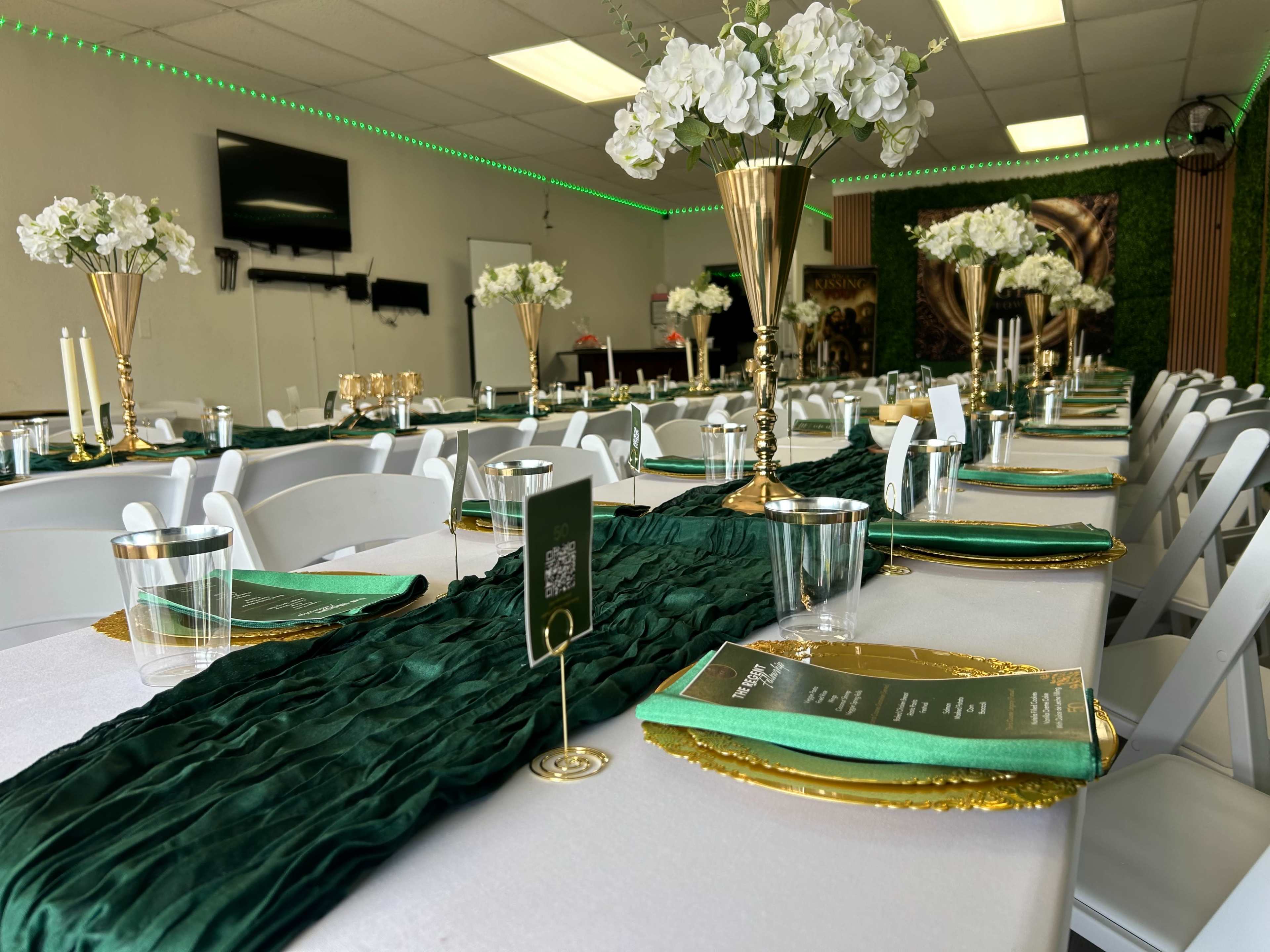 The image shows a decorated dining area with a long table set with green and gold tableware, flowers in vases, and candles, ready for an event.