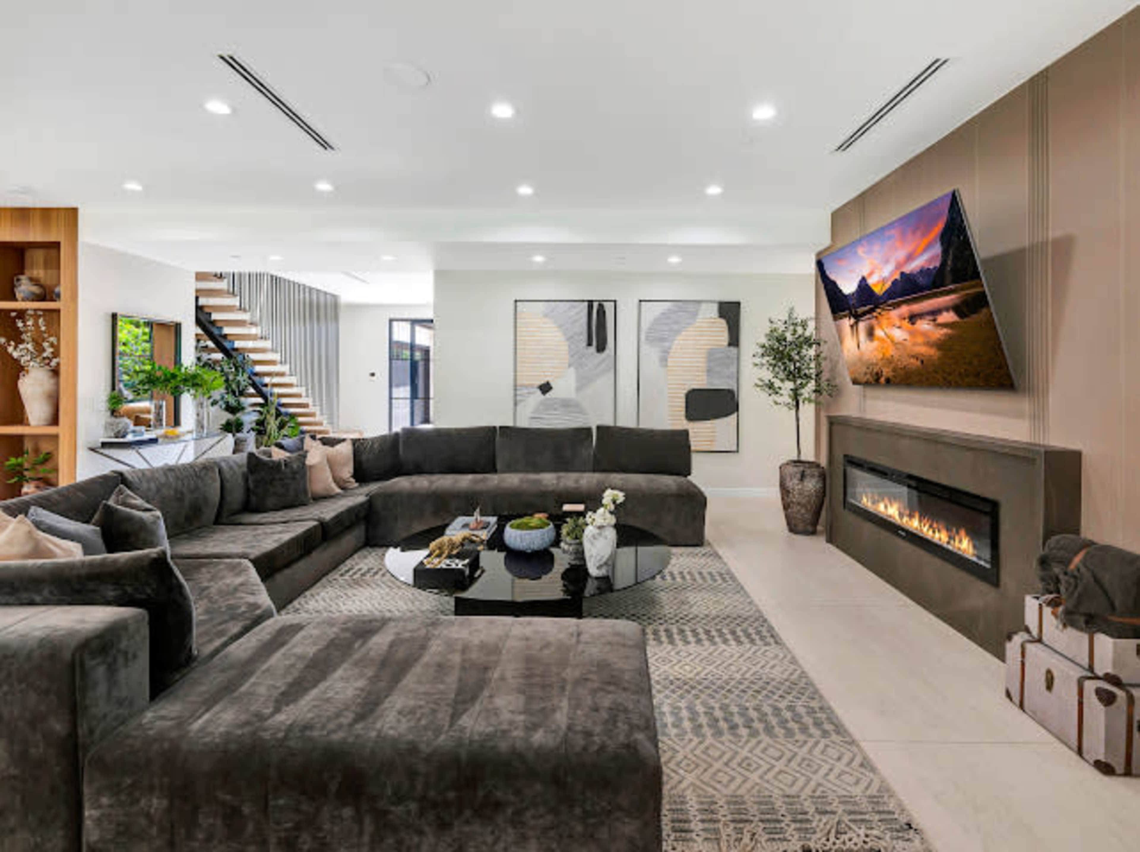 The image shows a modern living room featuring a large sectional sofa, a flat-screen TV mounted on a wall, a decorative fireplace, and a coffee table surrounded by various decorative items.