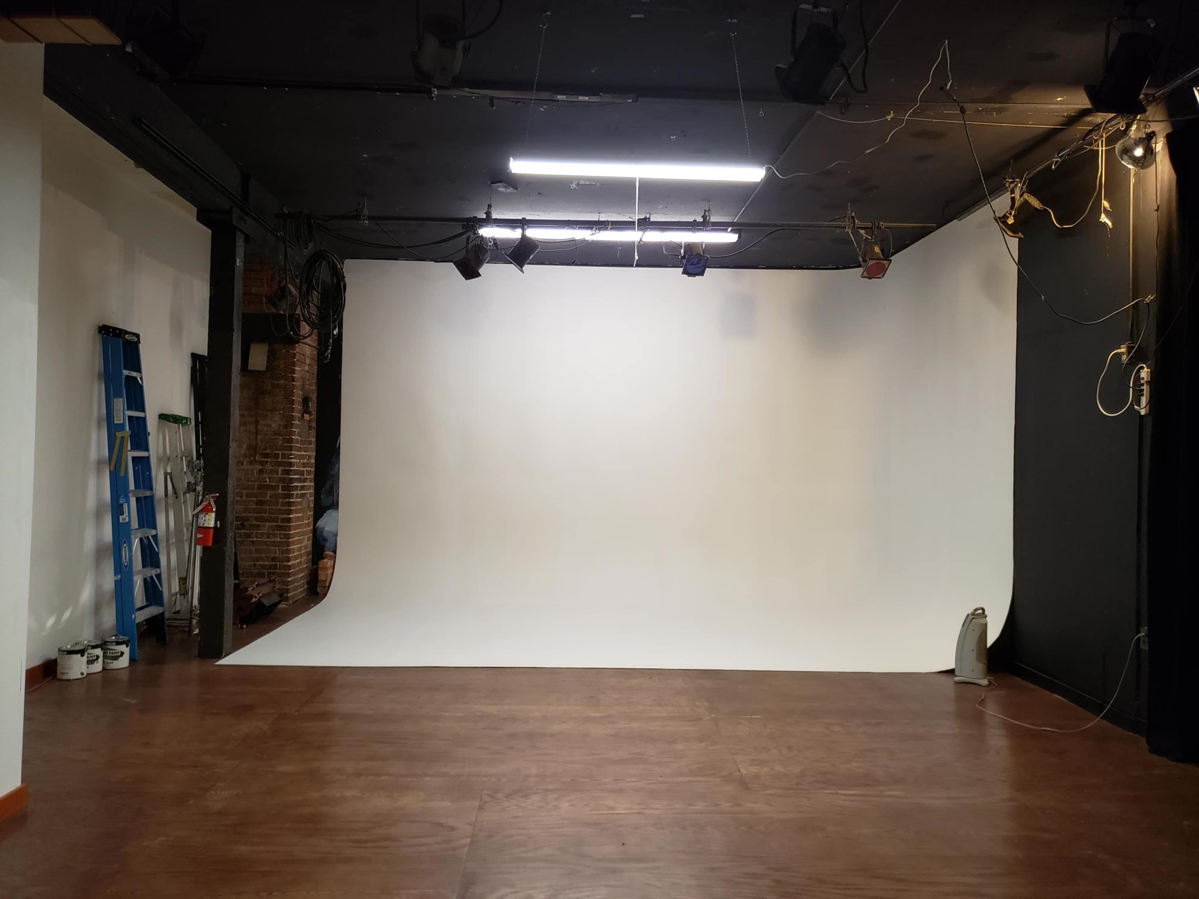 A photography studio with a white backdrop, wooden floor, and various lighting equipment.