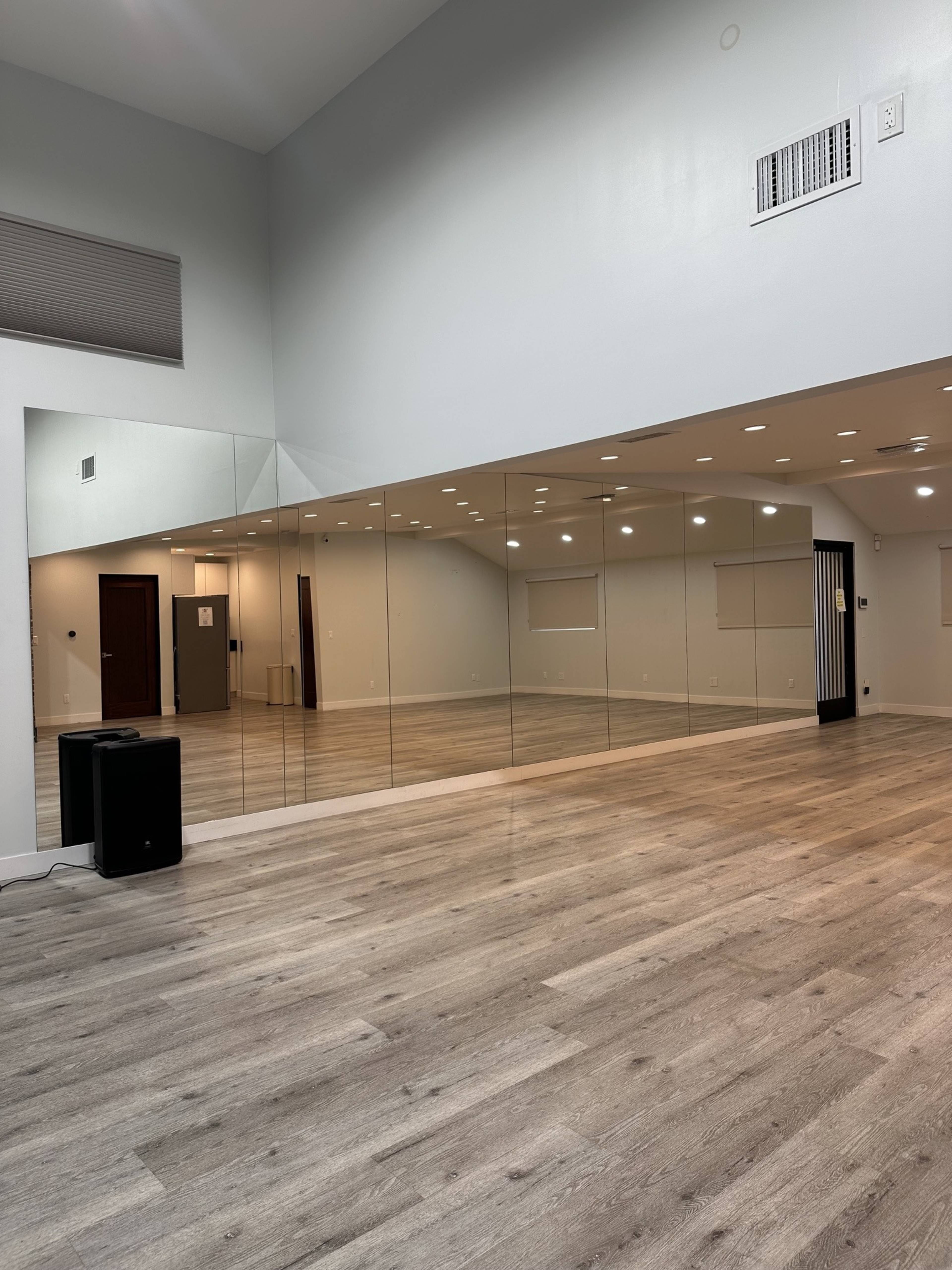 The image shows a spacious, well-lit room with a mirrored wall and light-colored wooden flooring.