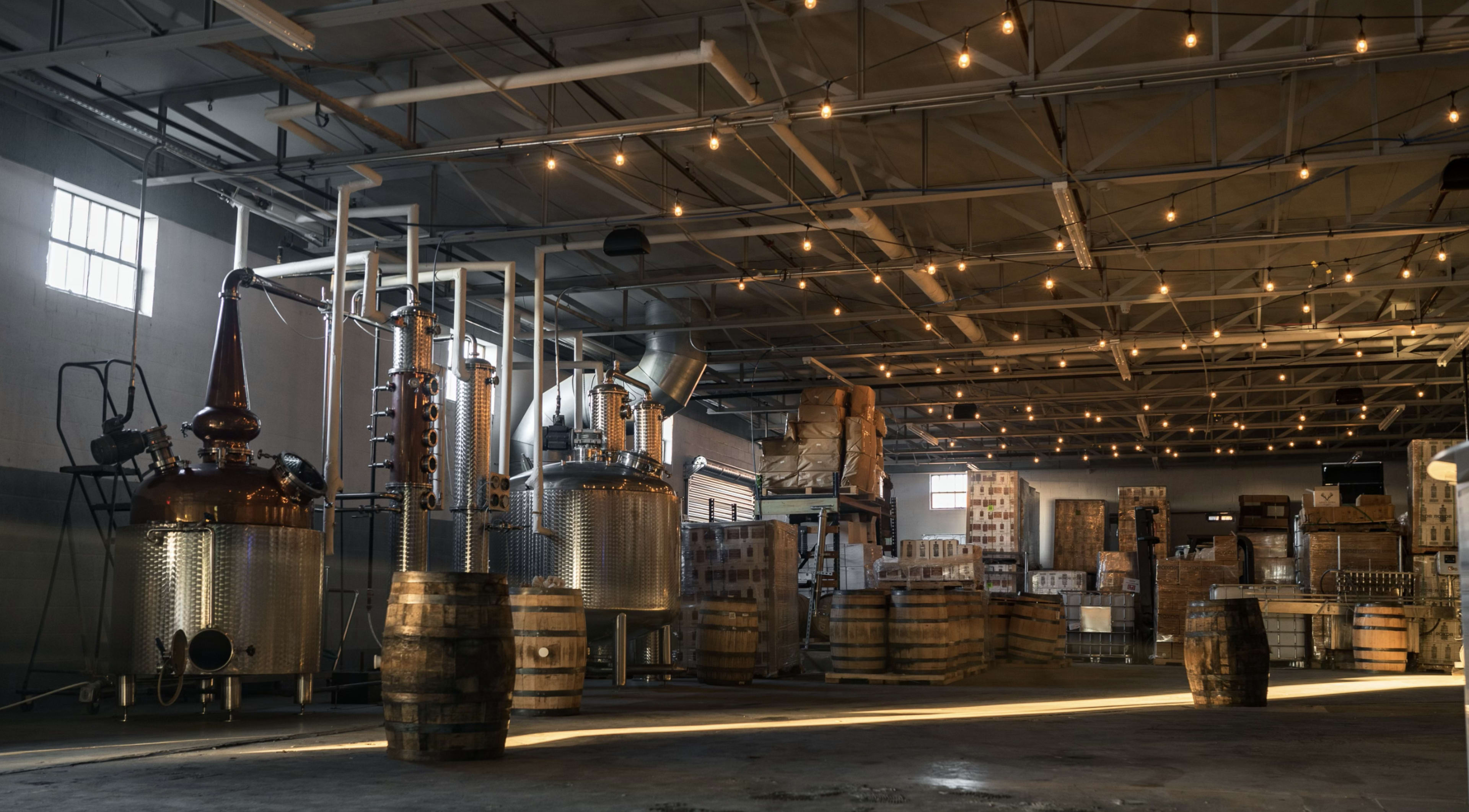 A distillery interior with metal distillation equipment, wooden barrels, and stacked boxes illuminated by string lights.