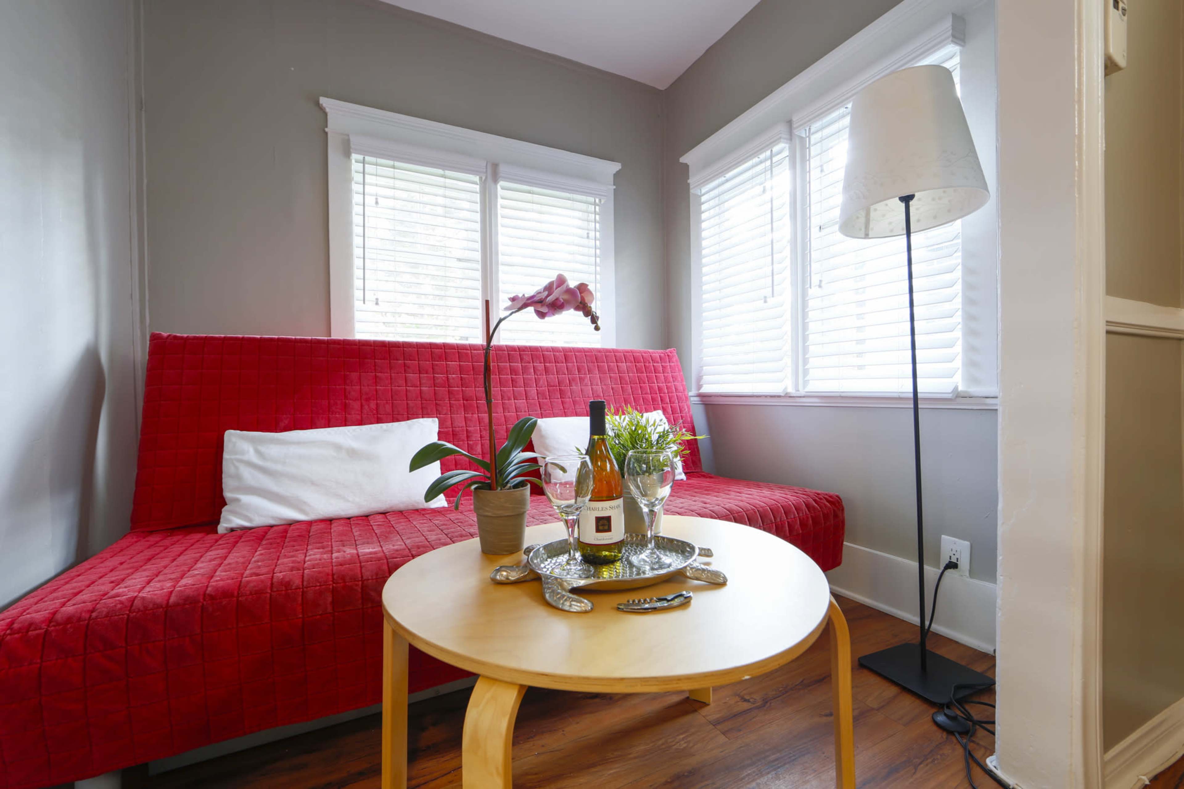 A red sofa with white pillows sits near a table with plants and decorative items, illuminated by natural light from the windows.