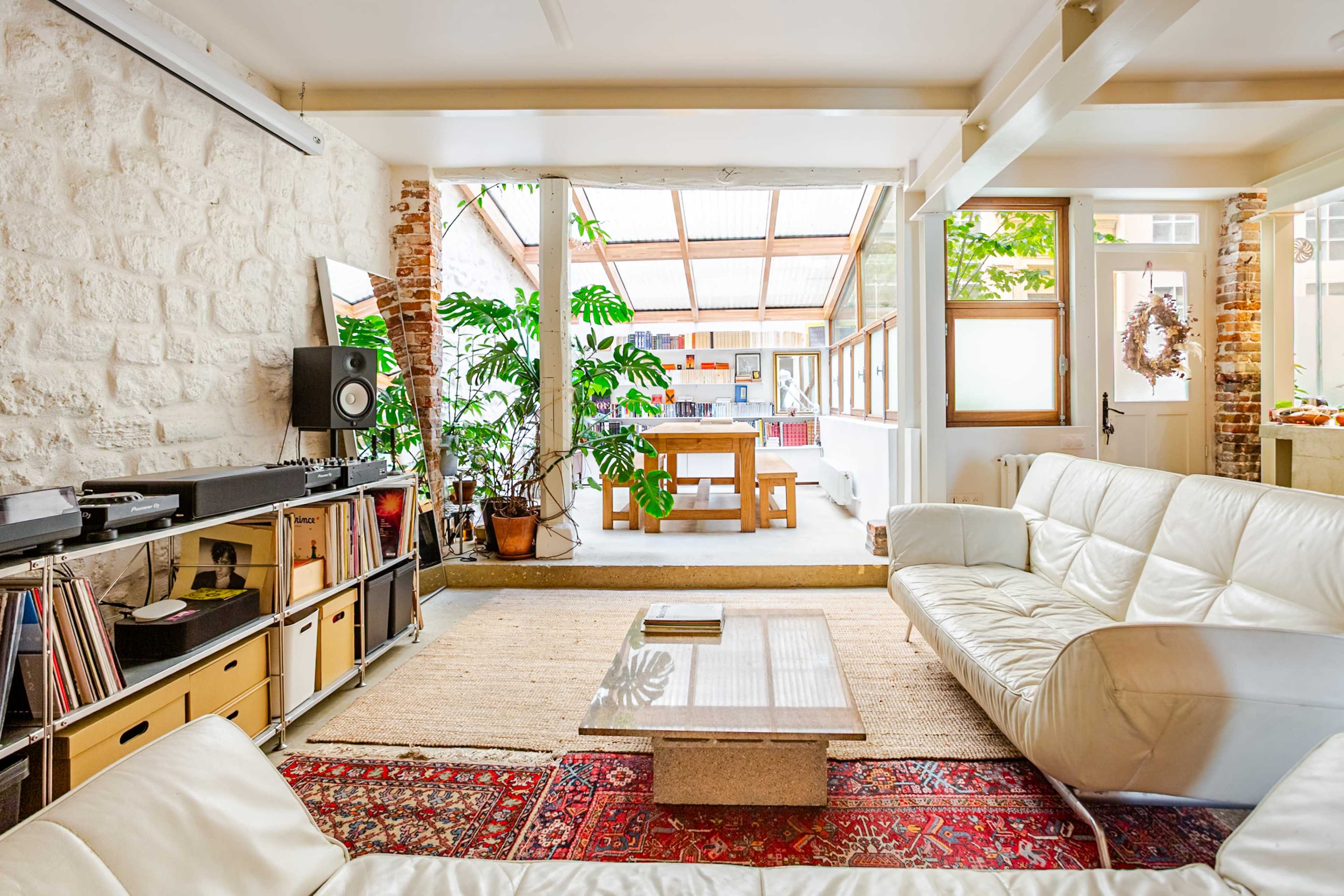 A modern living room features a white leather sofa, wooden dining table, large windows, and indoor plants against a backdrop of exposed brick walls.
