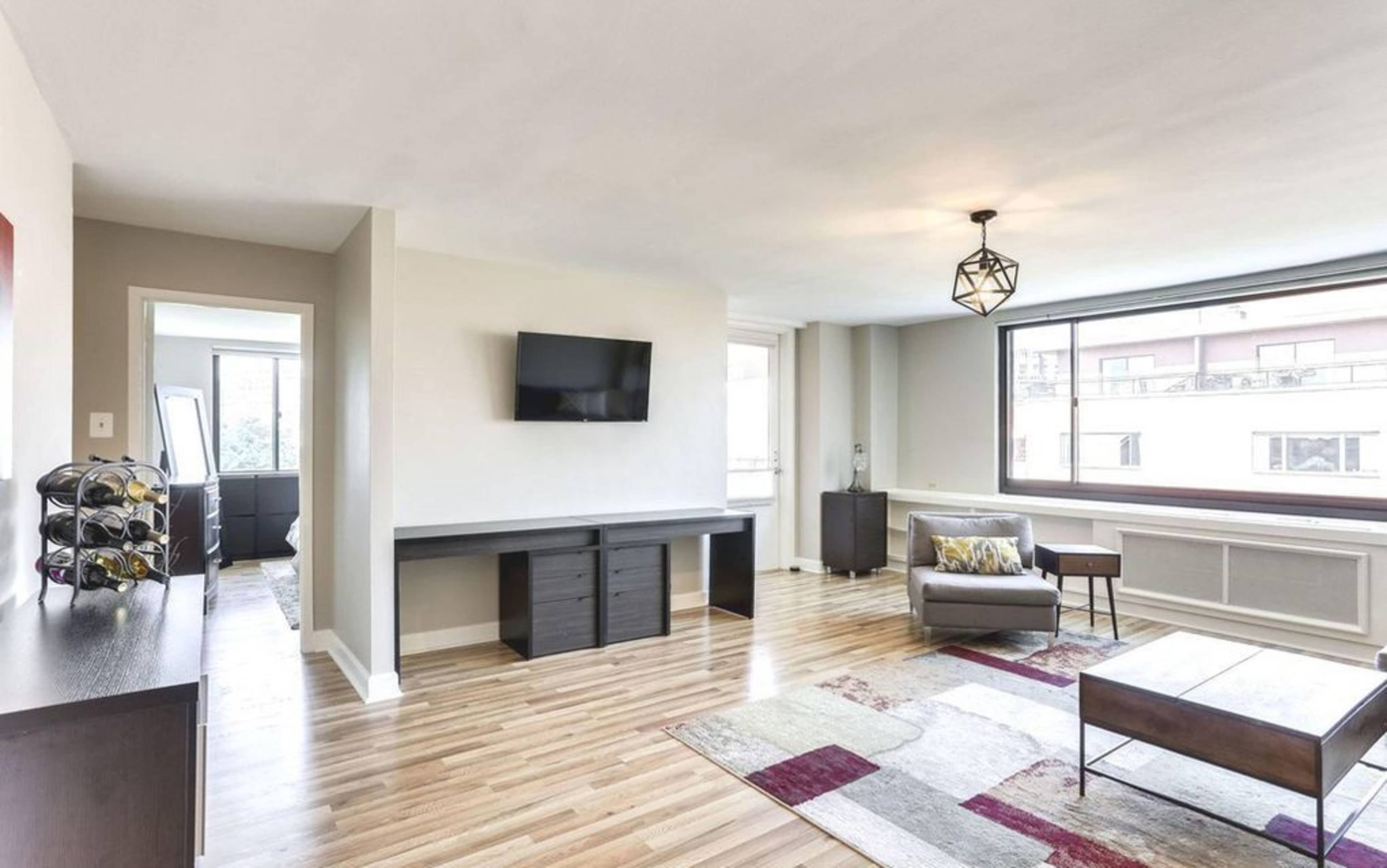 A modern, spacious living room with light hardwood floors, a wall-mounted TV, and a large window providing natural light.