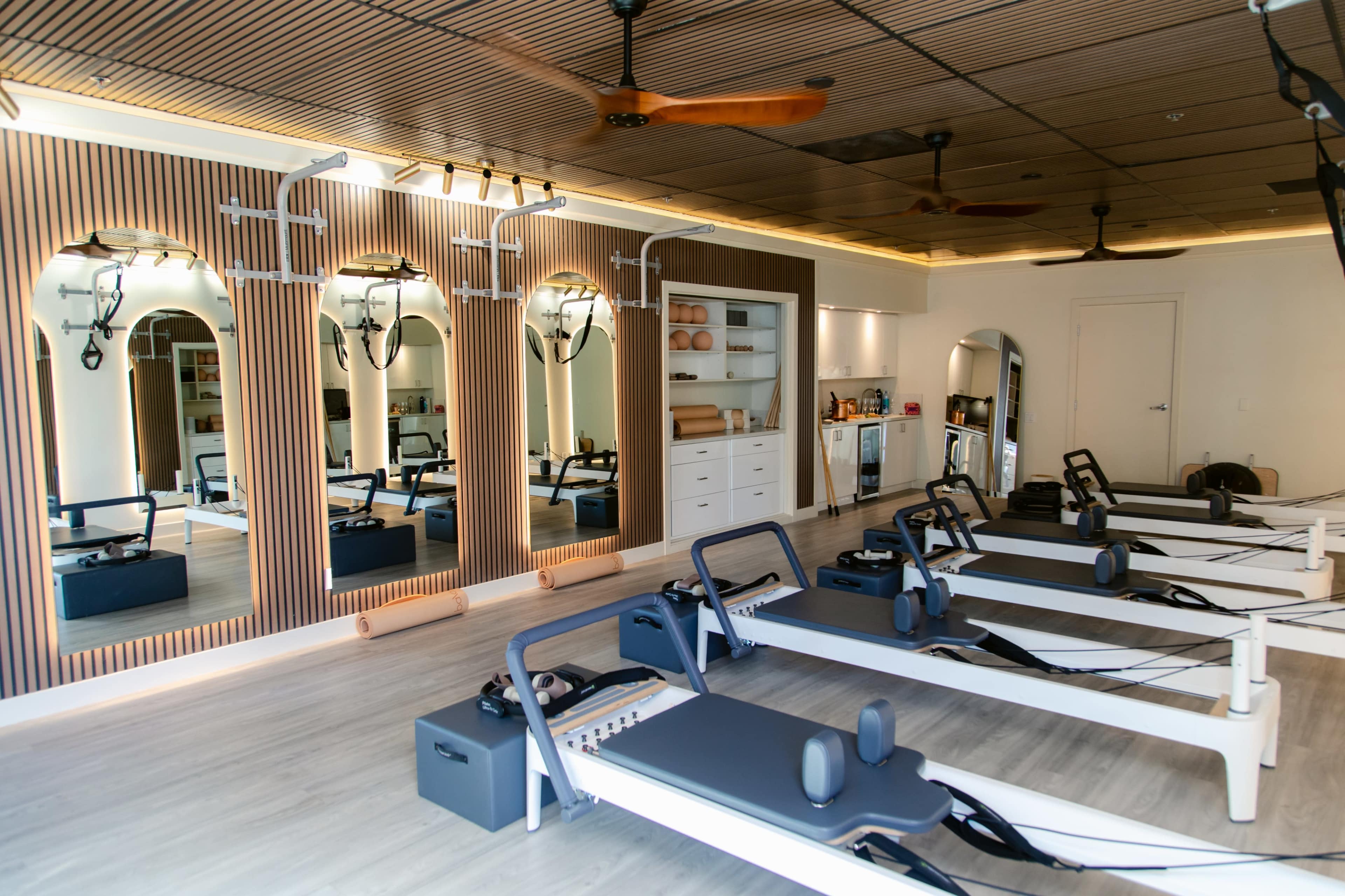 The image shows a Pilates studio with several reformer machines arranged in front of a wall of mirrors and a neatly organized storage area.