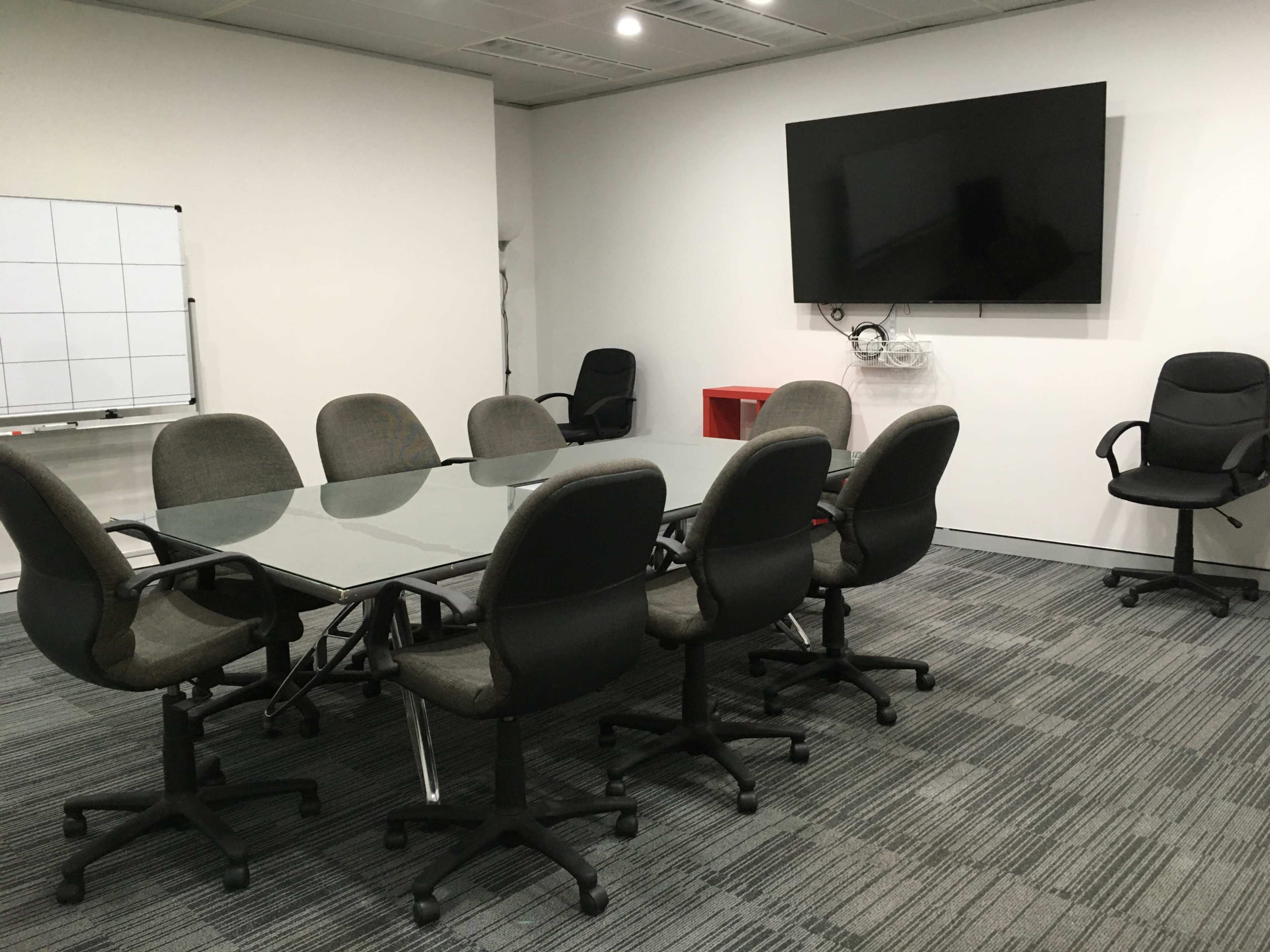 A conference room with a glass-topped table surrounded by eight upholstered chairs, a whiteboard, and a wall-mounted television.