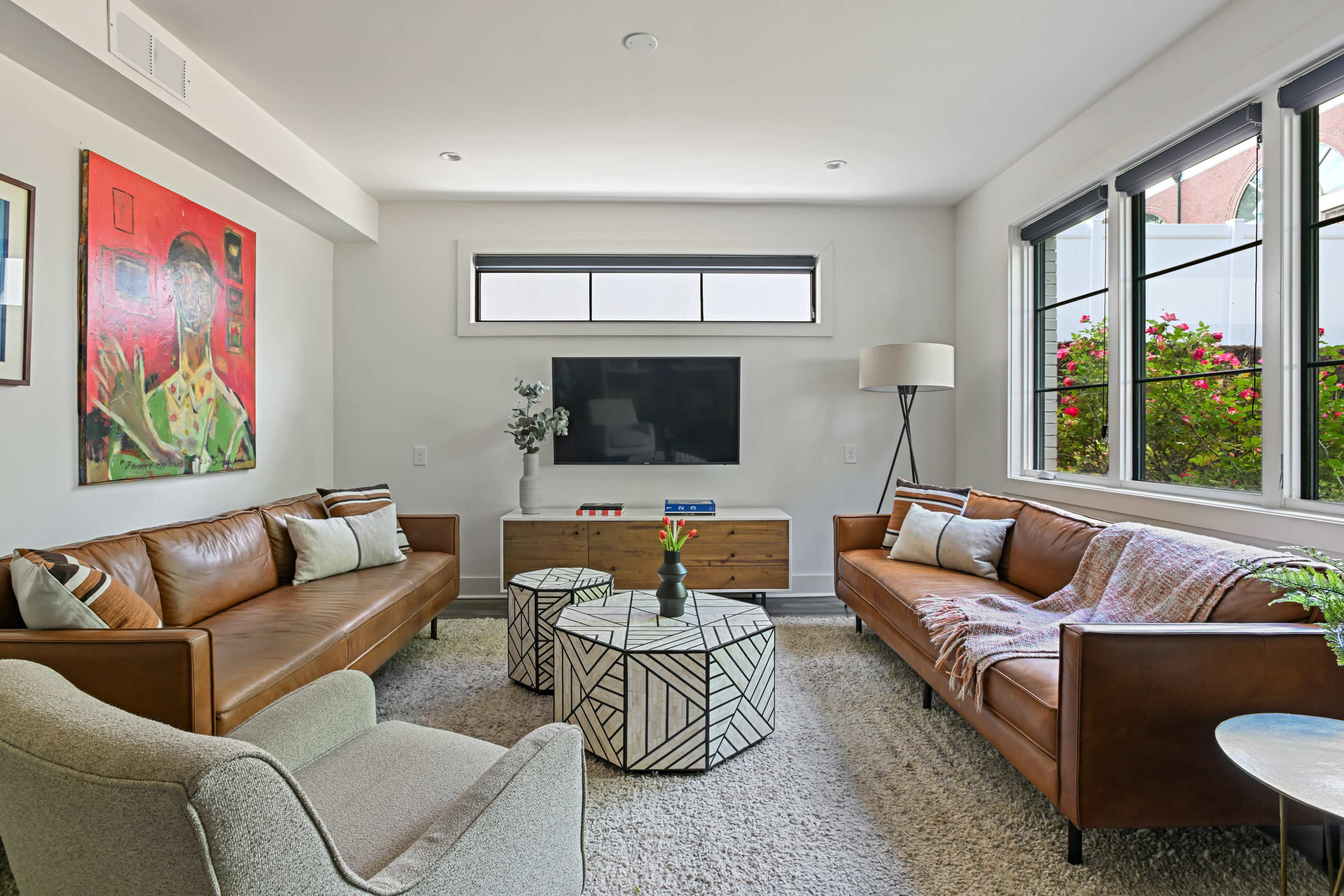 A modern living room features two leather sofas, a patterned coffee table, a television mounted on the wall, and large windows with a view of greenery outside.