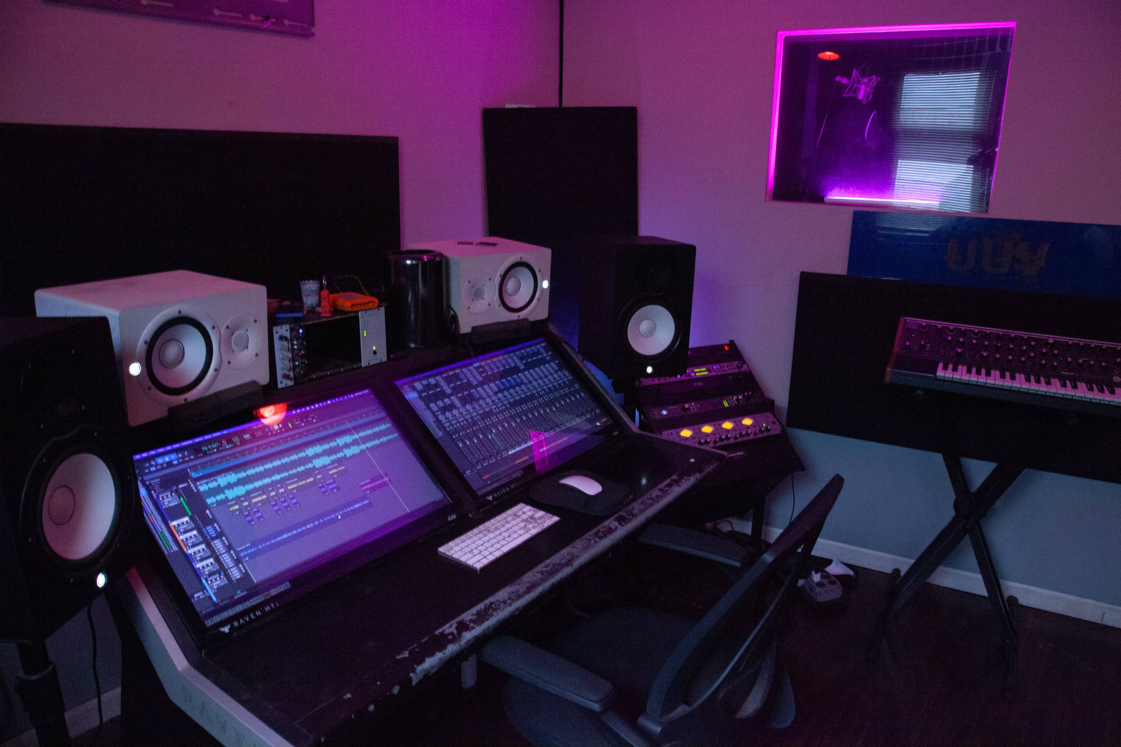 The image shows a recording studio with a large mixing console, monitor speakers, and a synthesizer against a backdrop of purple lighting.