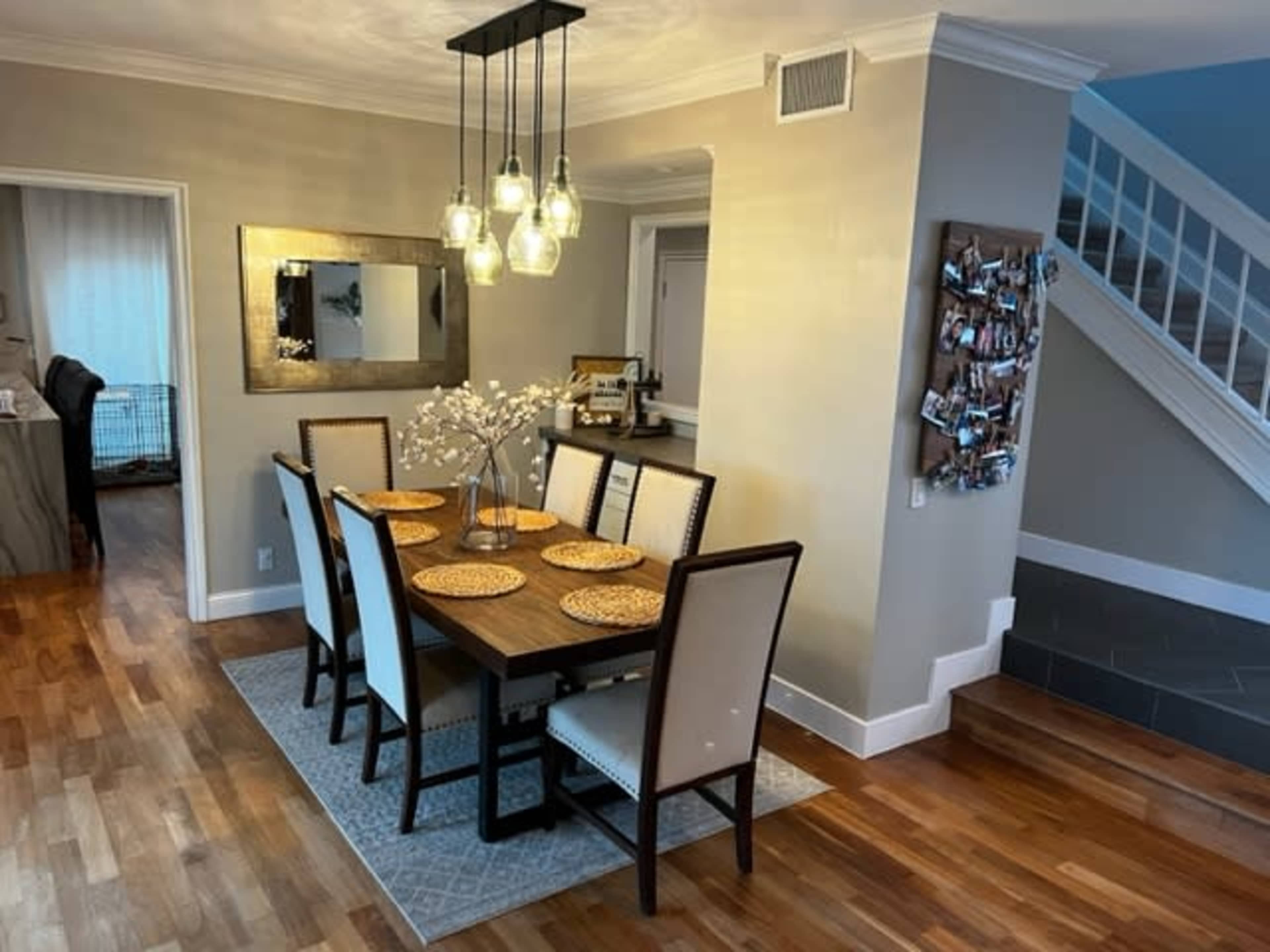 The image shows a dining area with a wooden table set for eight and a modern chandelier, surrounded by light-colored walls and hardwood flooring.