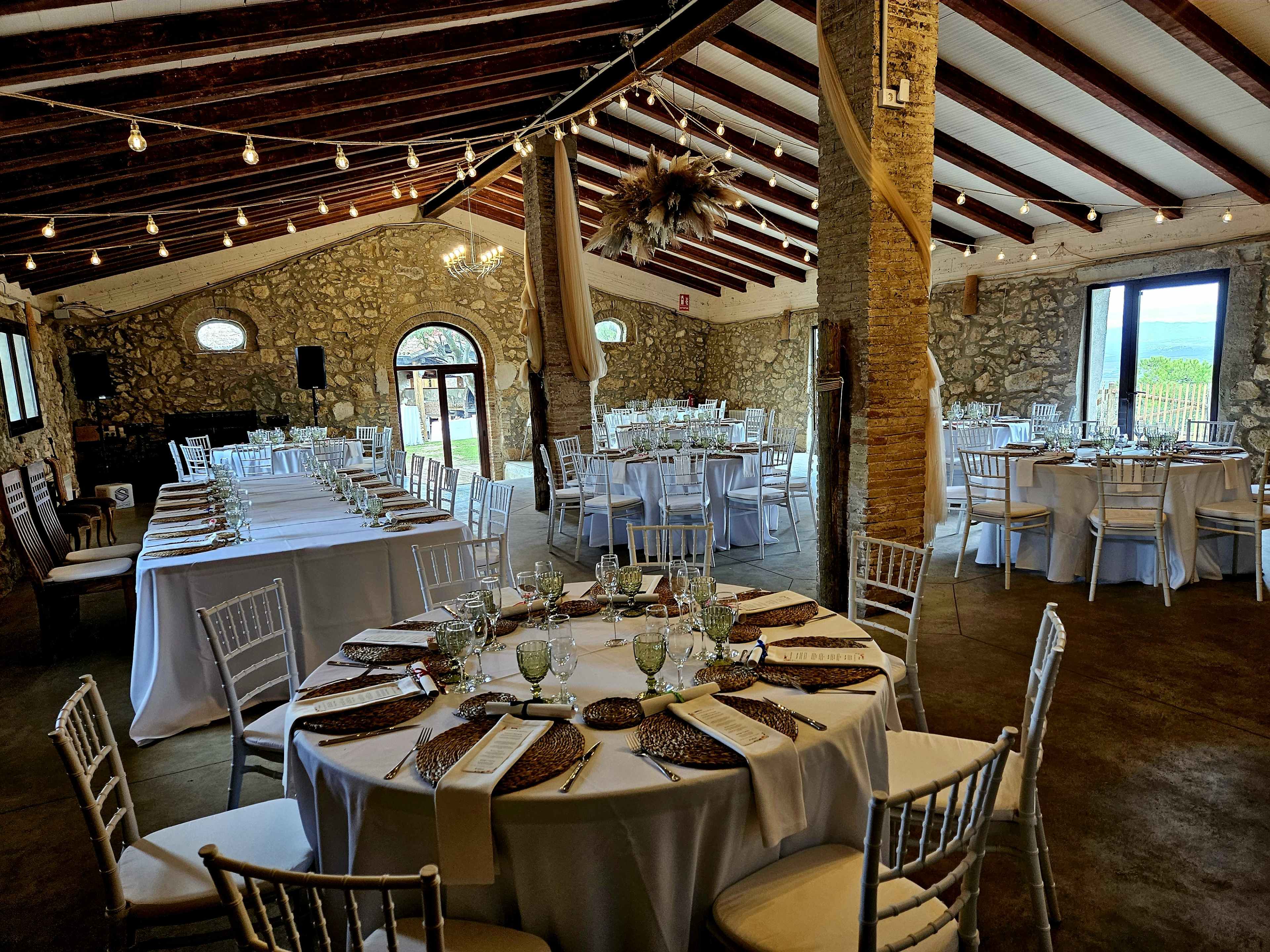 A spacious venue with stone walls features elegantly set round and rectangular dining tables, decorated with white linens and glassware, under wooden beams and string lights.