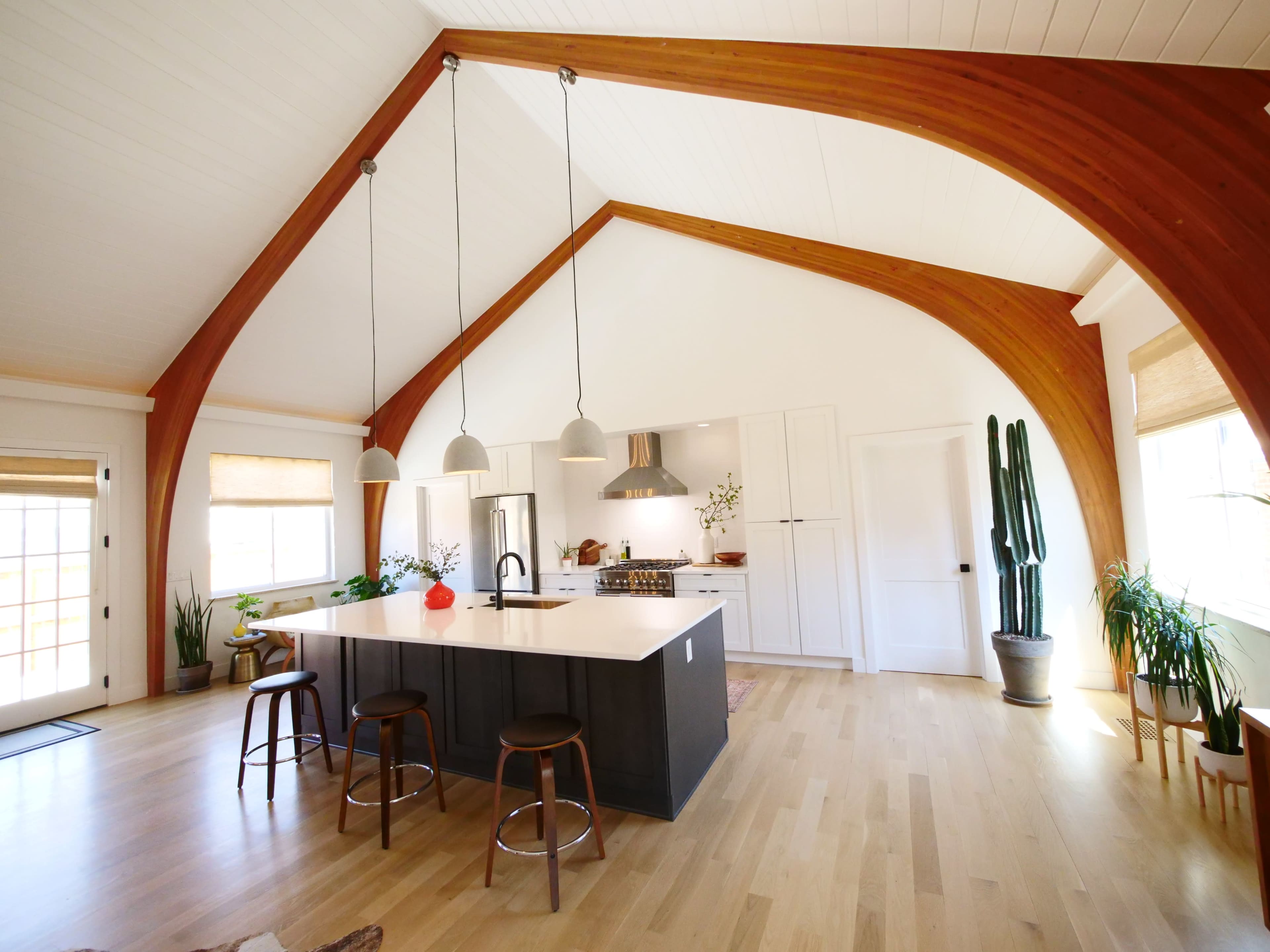 A modern kitchen with an island, wooden beams, and large windows, featuring a variety of plants and pendant lighting.