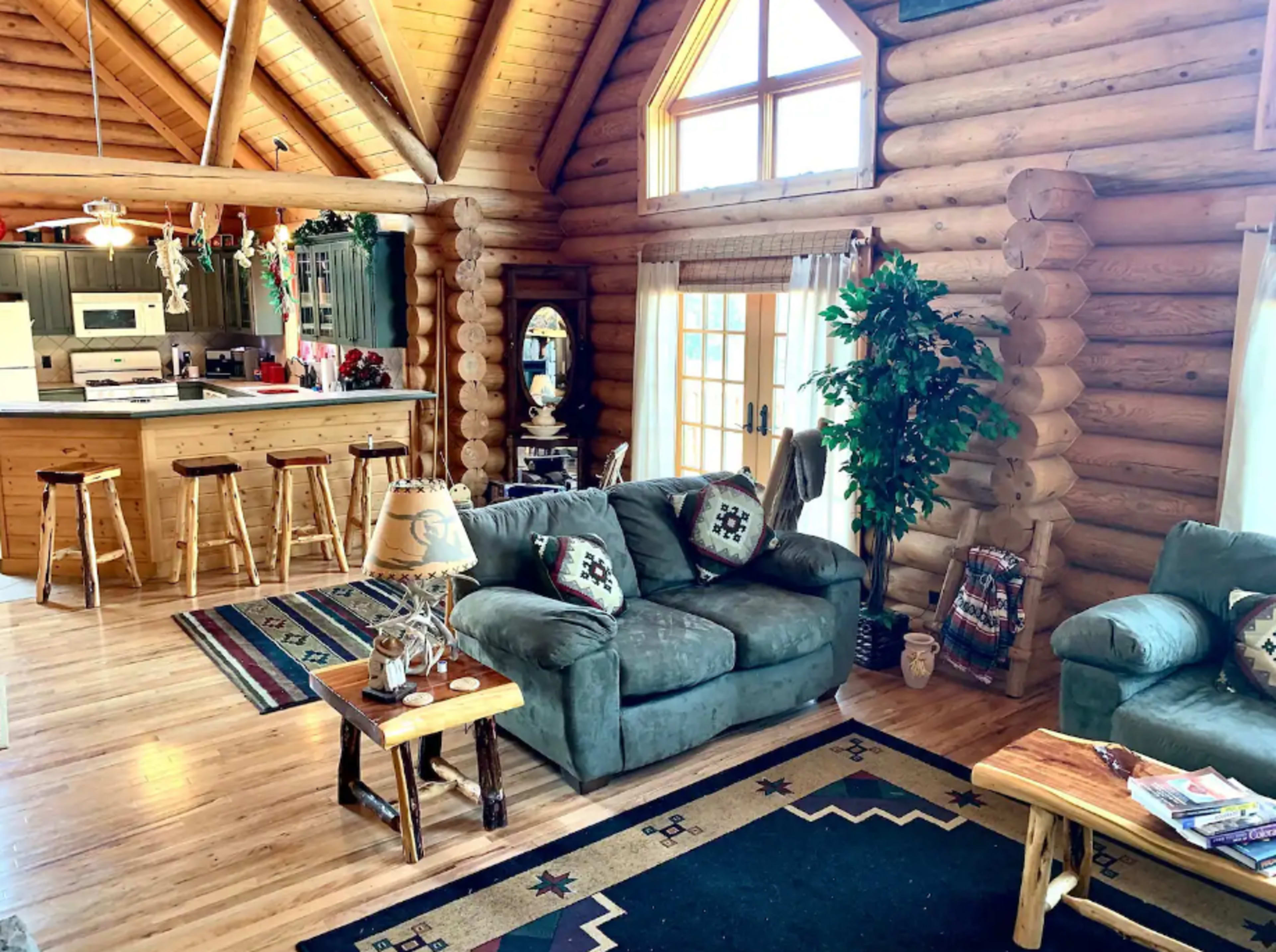 The image shows a spacious log cabin interior featuring a living area with two sofas, a coffee table, and a kitchen with bar stools in the background.