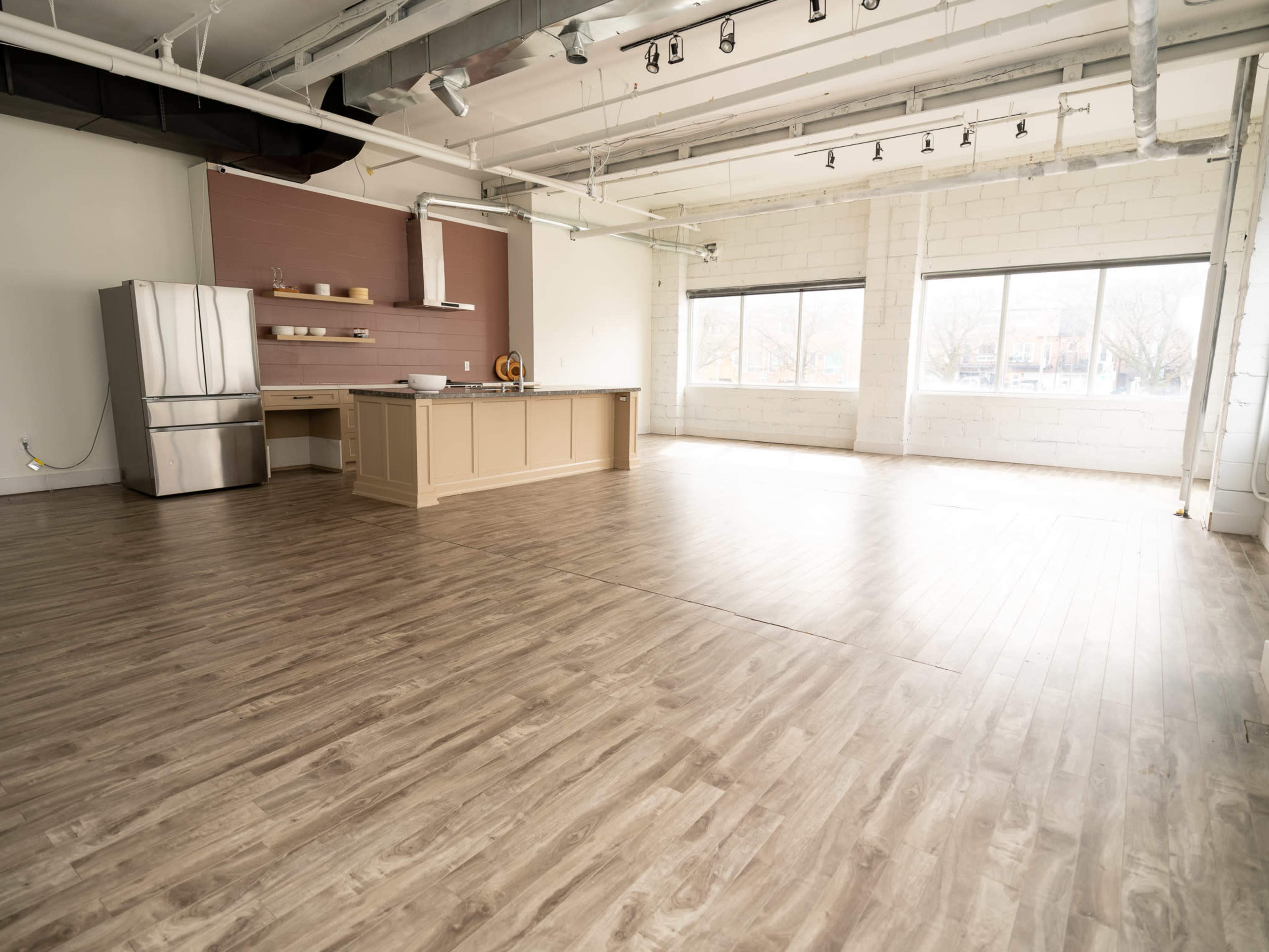 The image shows a spacious, empty room with light wood flooring, large windows, and a central kitchen island with a stainless steel refrigerator nearby.