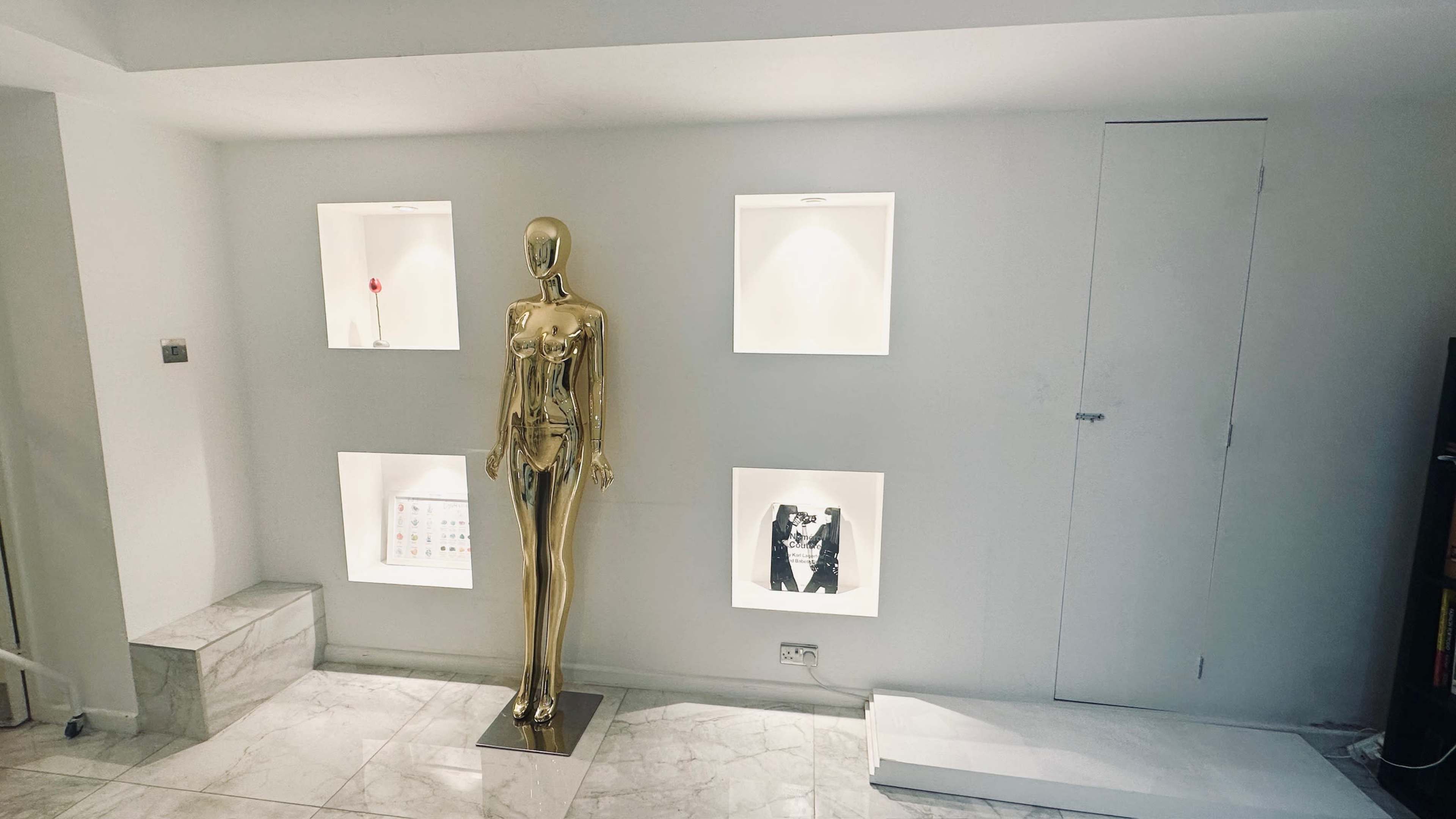 A gold mannequin stands in a minimalist room with white walls, flanked by three illuminated square niches containing decorative items.