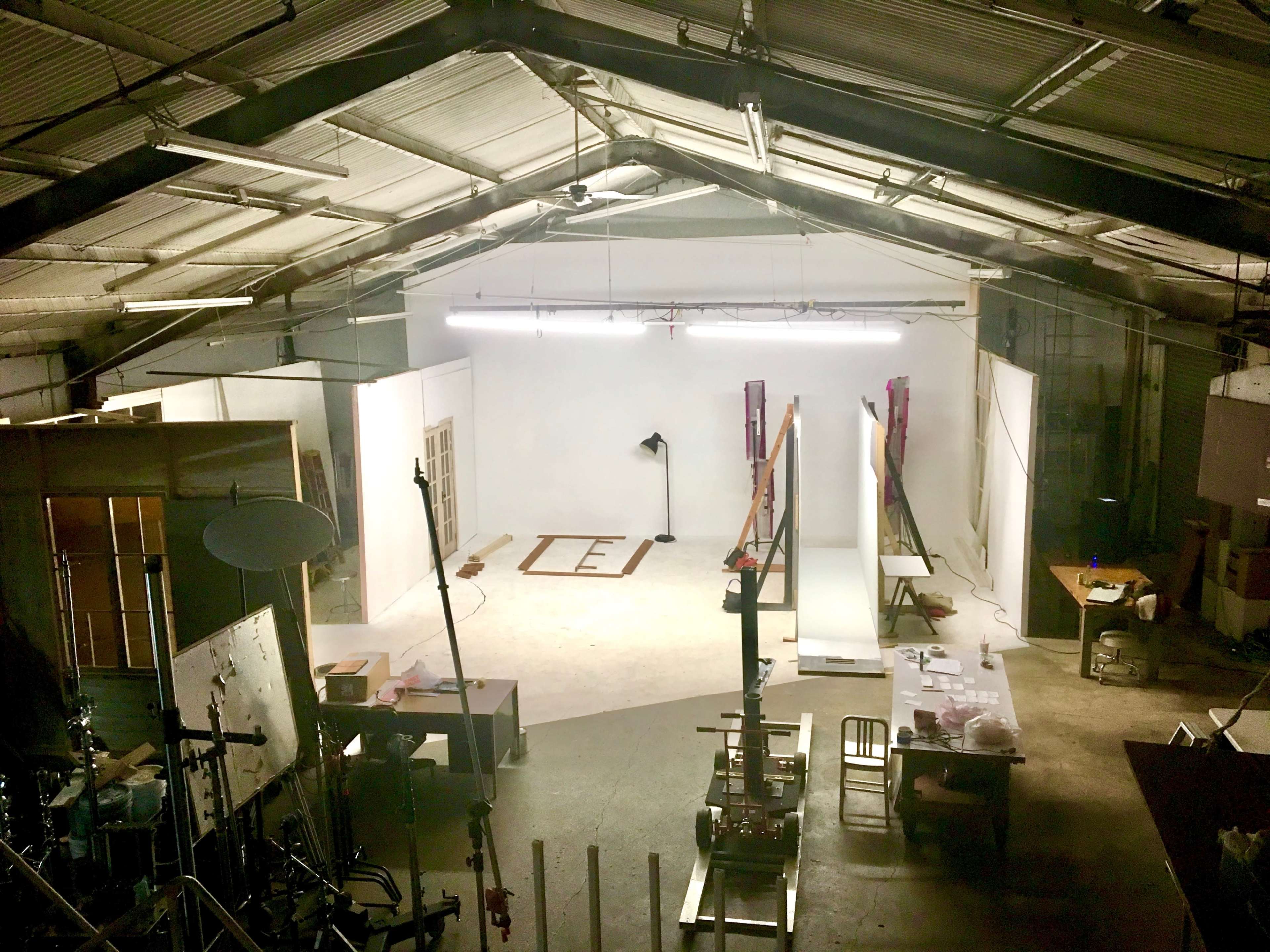The image shows an industrial-style studio space with a white backdrop, various equipment, and scattered furniture, illuminated by overhead lights.
